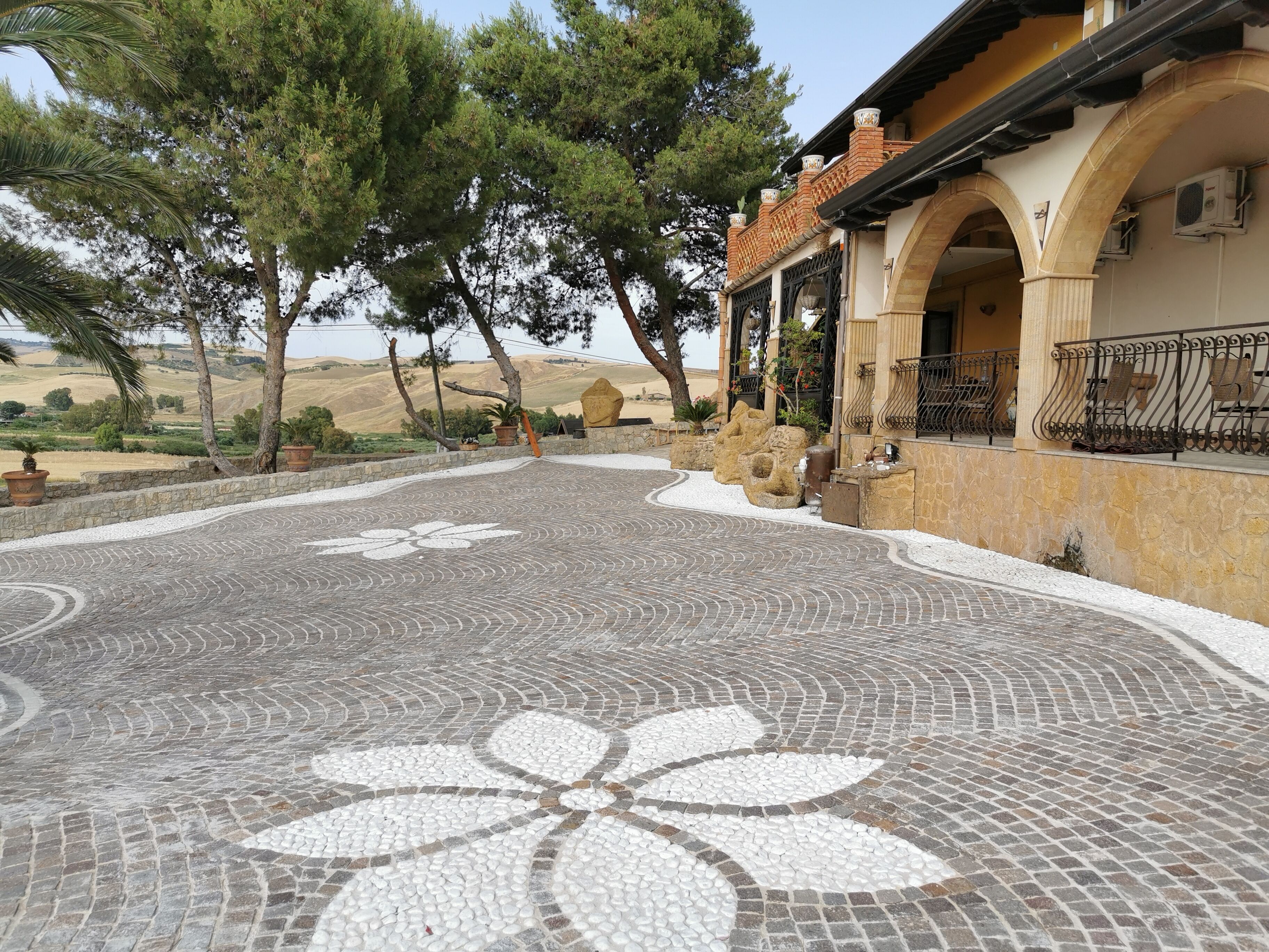 Une cour avec un sol en mosaïque, des murs en pierre et un bâtiment doté d’arcades et d’un espace extérieur aménagé pour s’asseoir.
