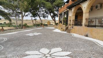 Une cour avec un sol en mosaïque, des murs en pierre et un bâtiment doté d’arcades et d’un espace extérieur aménagé pour s’asseoir.
