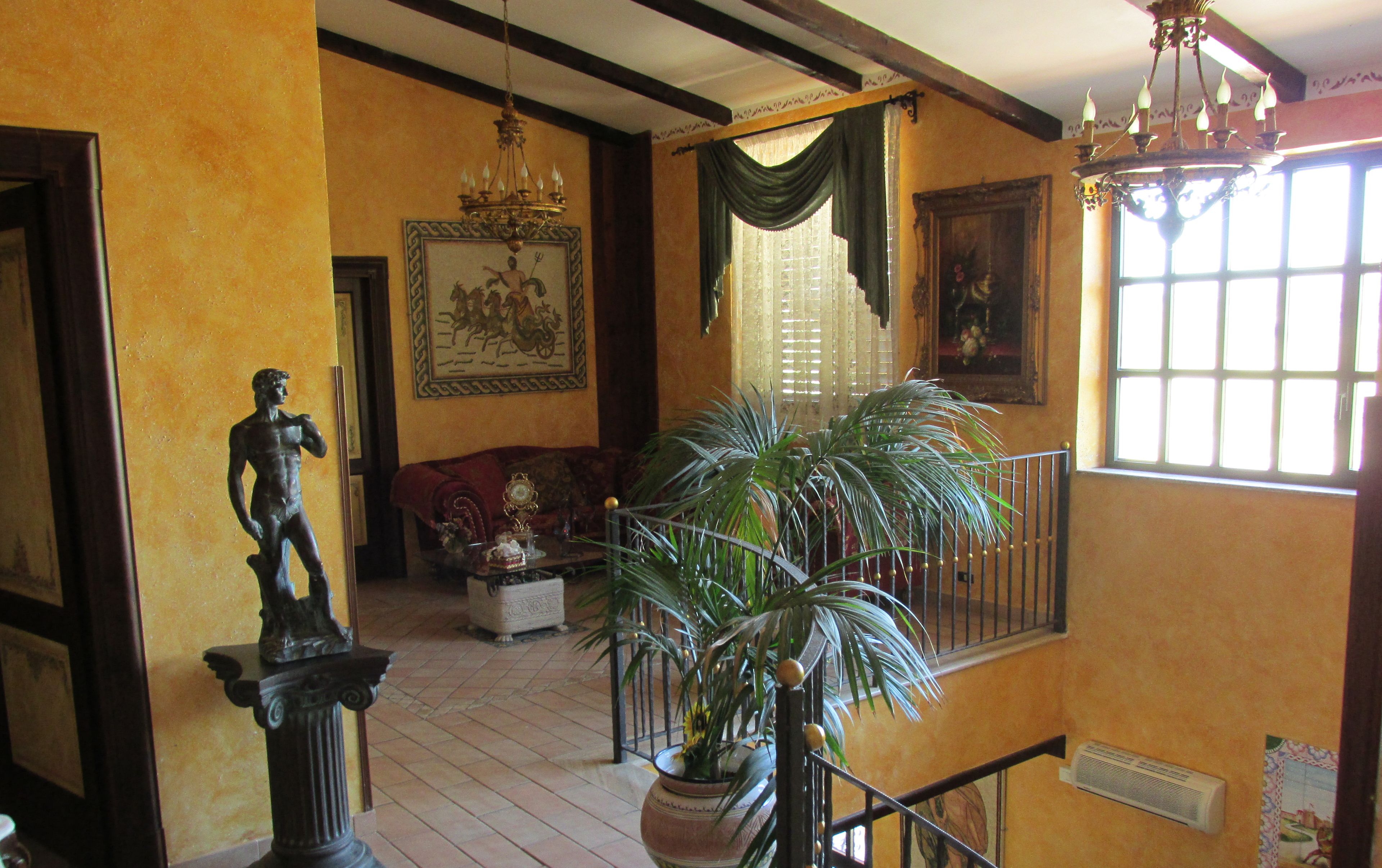 Um quarto com piso de madeira, uma estátua, um sofá, um lustre e uma janela grande com cortinas.