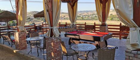 An outdoor patio area with a pool table, chairs, and a view of the countryside.