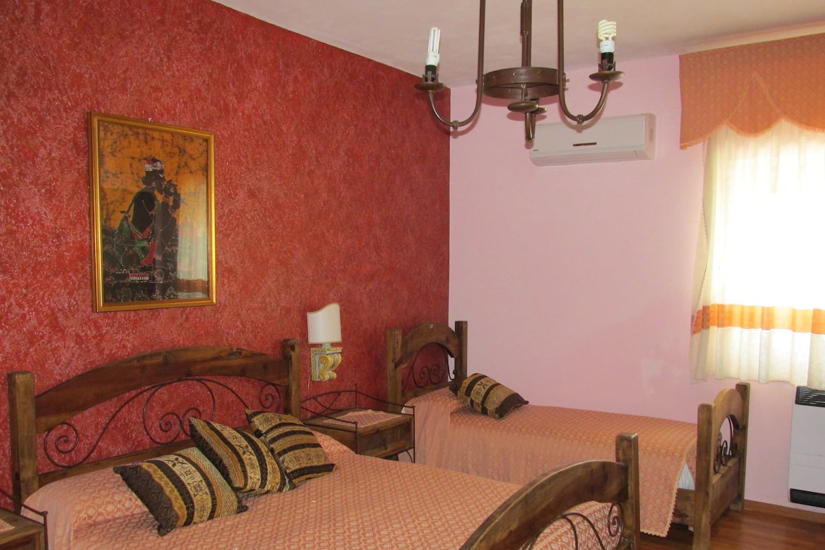 Un dormitorio con dos camas de madera, un cuadro en la pared, una araña y una ventana con cortinas.