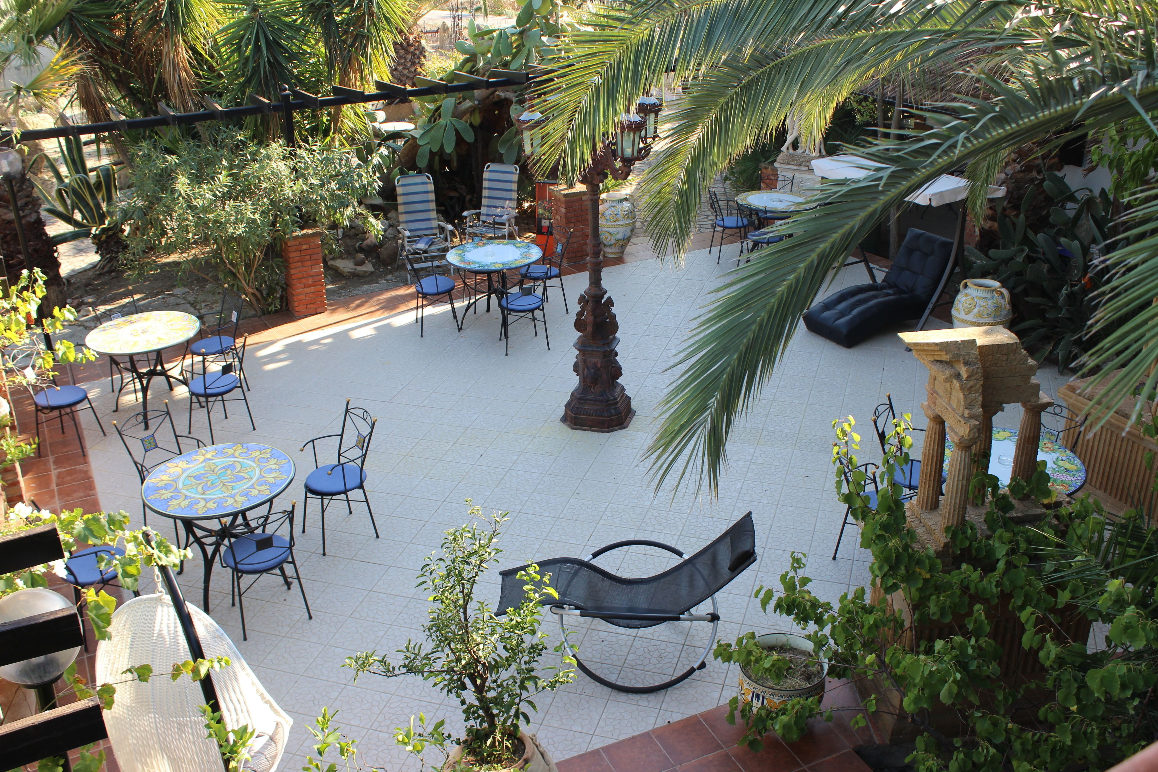 An outdoor patio with multiple tables and chairs, surrounded by various plants and a palm tree.