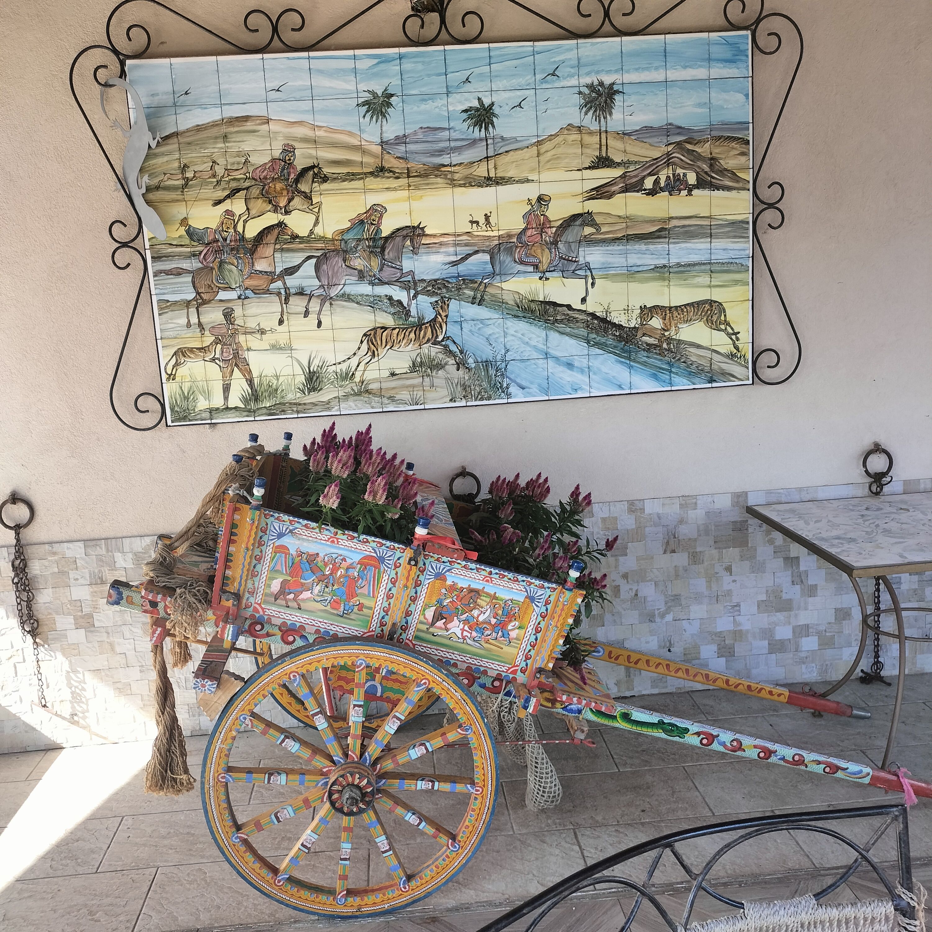 Un mosaico mural que muestra una escena desértica con camellos y jinetes, un carro decorativo con diseños coloridos y una pequeña mesa con estructura metálica.