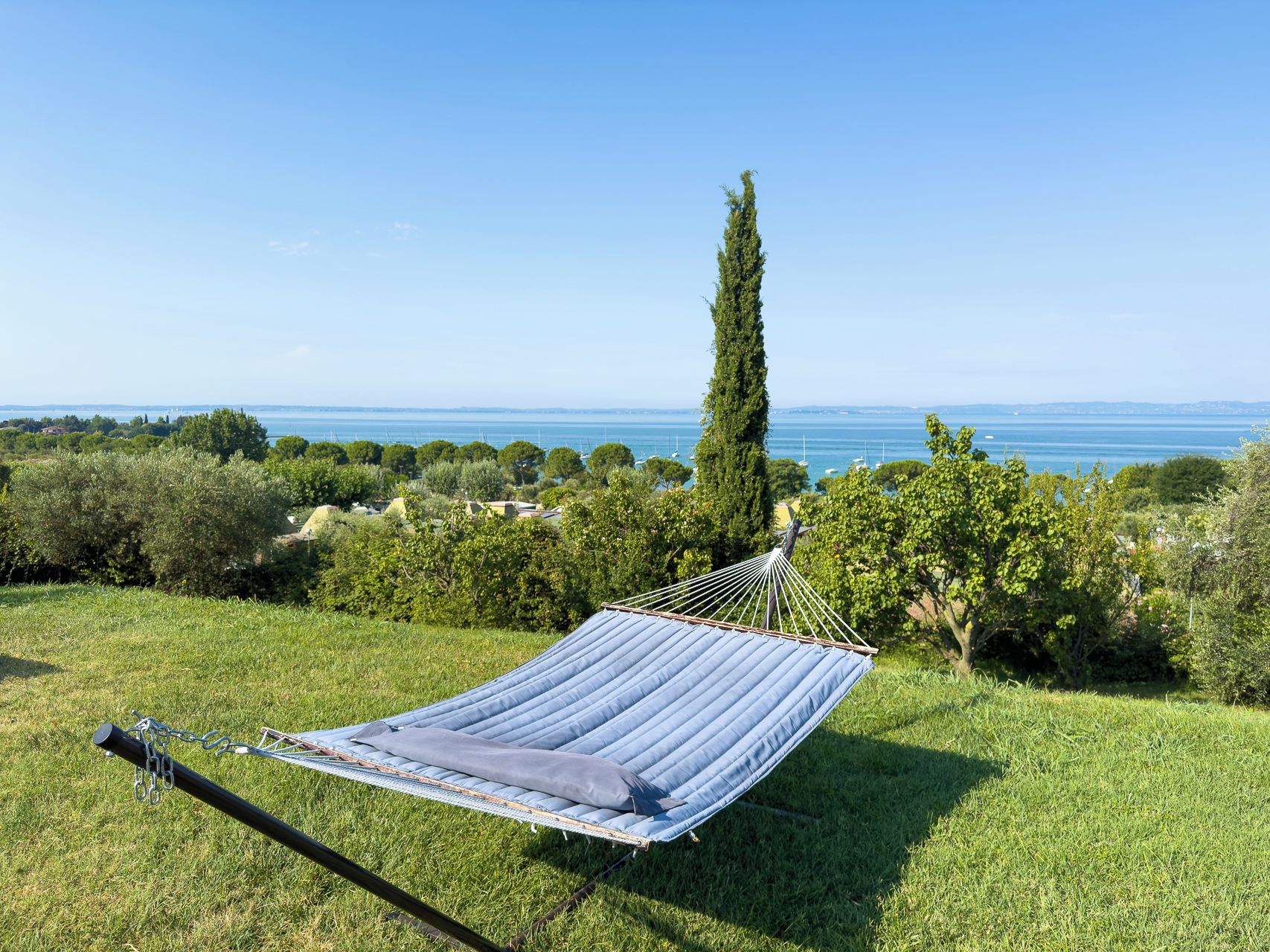 A hammock on a grassy hill overlooking a body of water with trees and buildings in the distance.