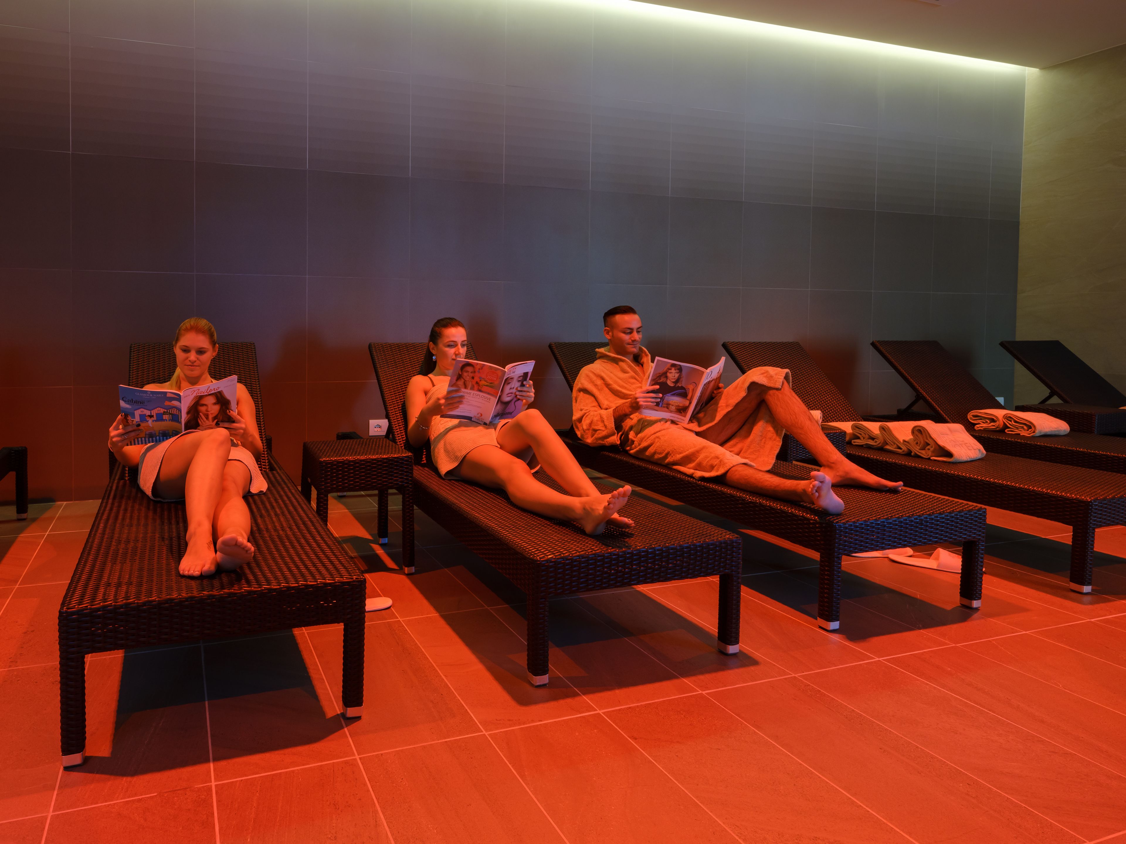 Trois personnes se détendent sur des fauteuils en osier, dans un cadre évoquant un spa, avec des murs carrelés et un sol rouge.