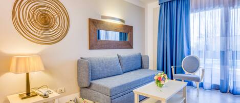 A modern living room with a sofa, a coffee table, a lamp, a phone, and a decorative mirror. There are blue curtains and a window with natural light.