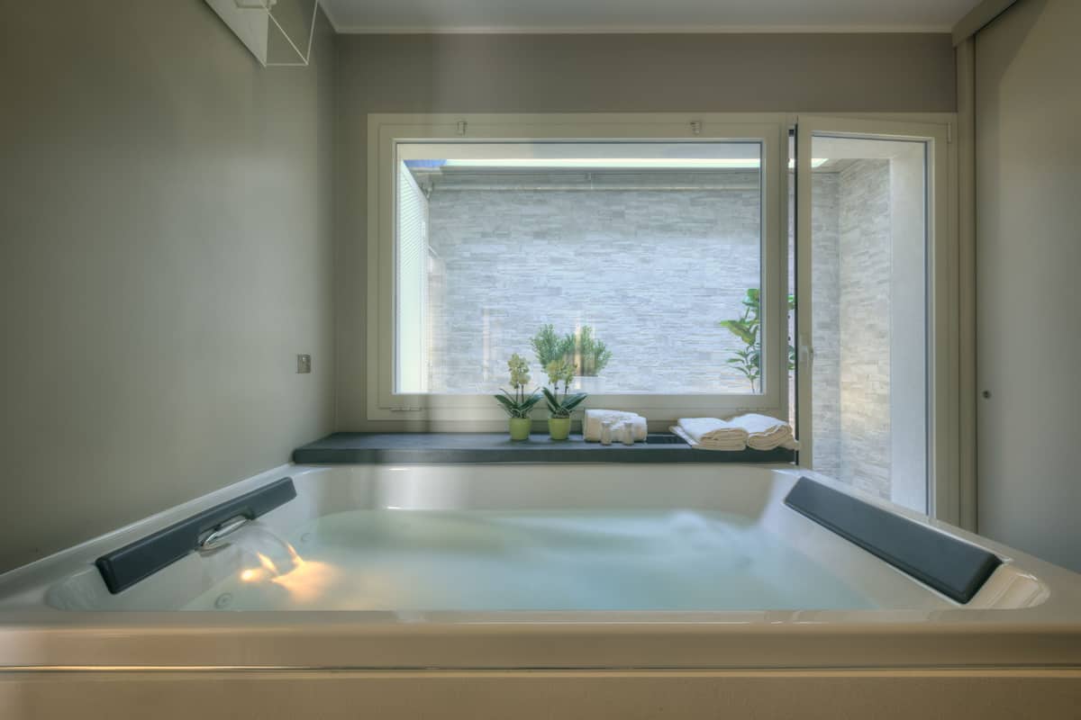 A spa room with a large bathtub, a window with a view of a brick wall and plants, and a shelf with towels and toiletries.