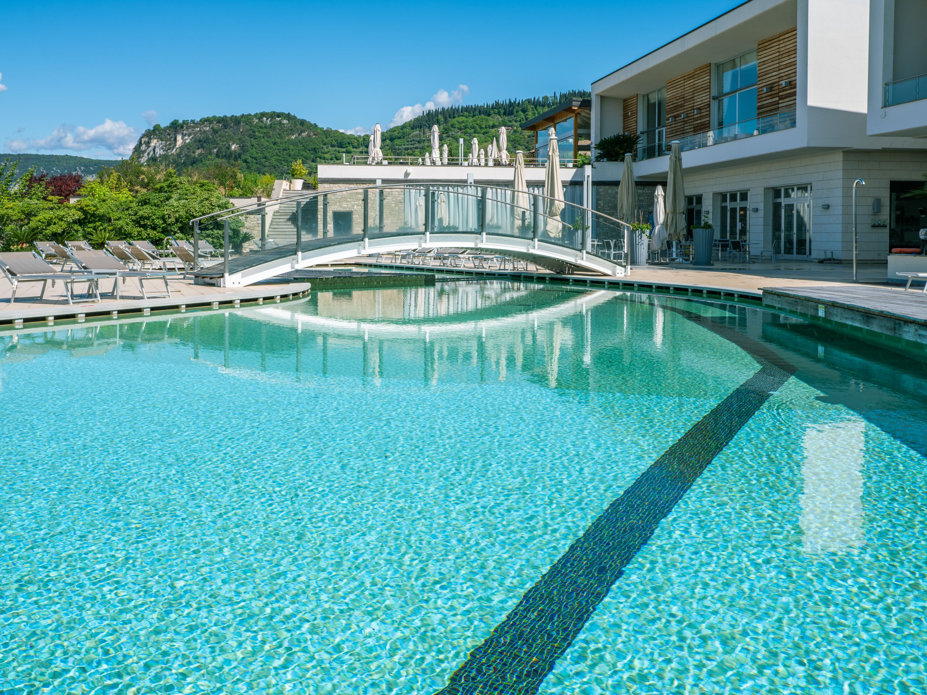 A clear blue pool with a glass bridge, lounge chairs, and a modern building with a mountain view in the background.