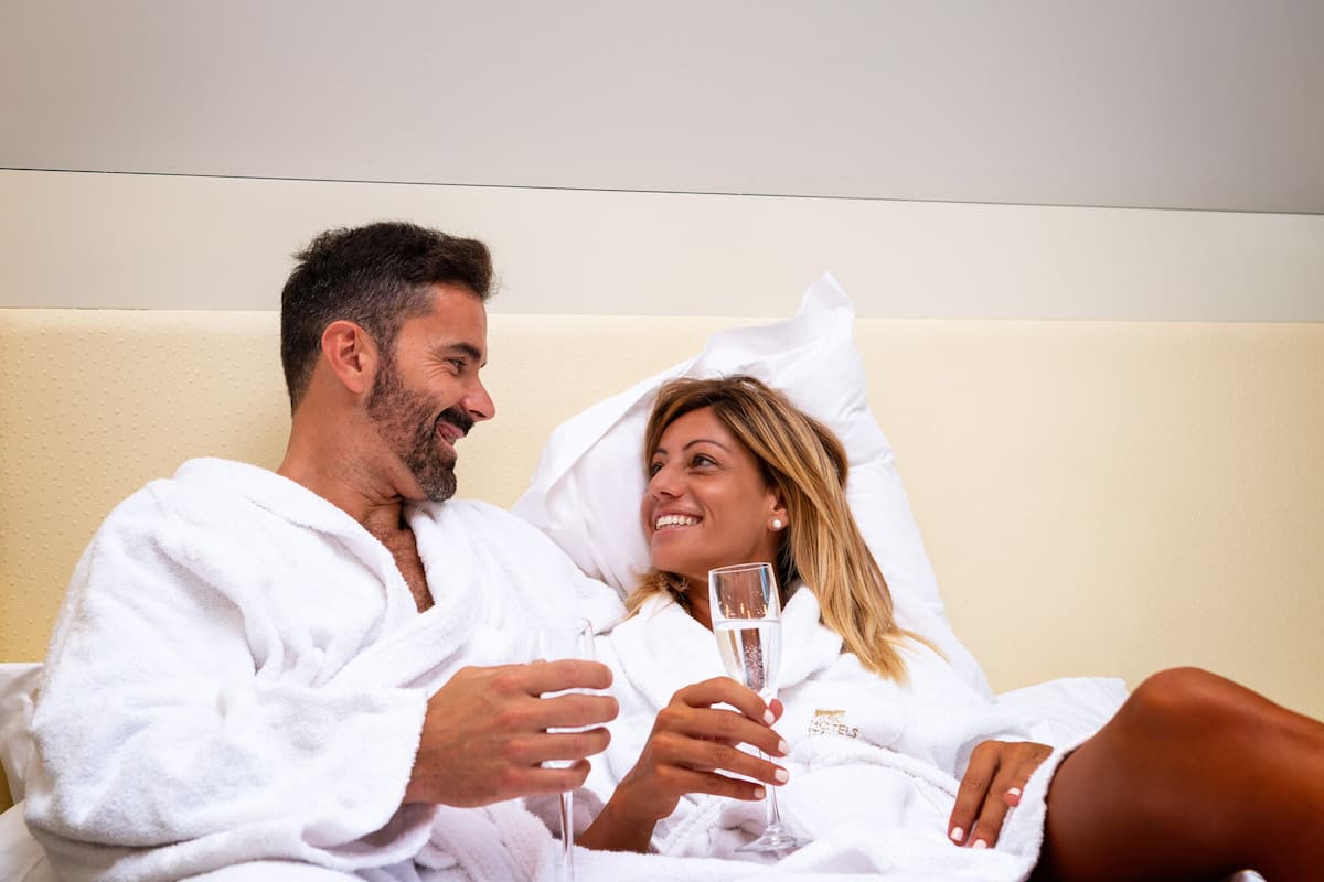 A couple in white bathrobes sitting on a bed with a champagne glass, in a room with a plain wall and a textured headboard.