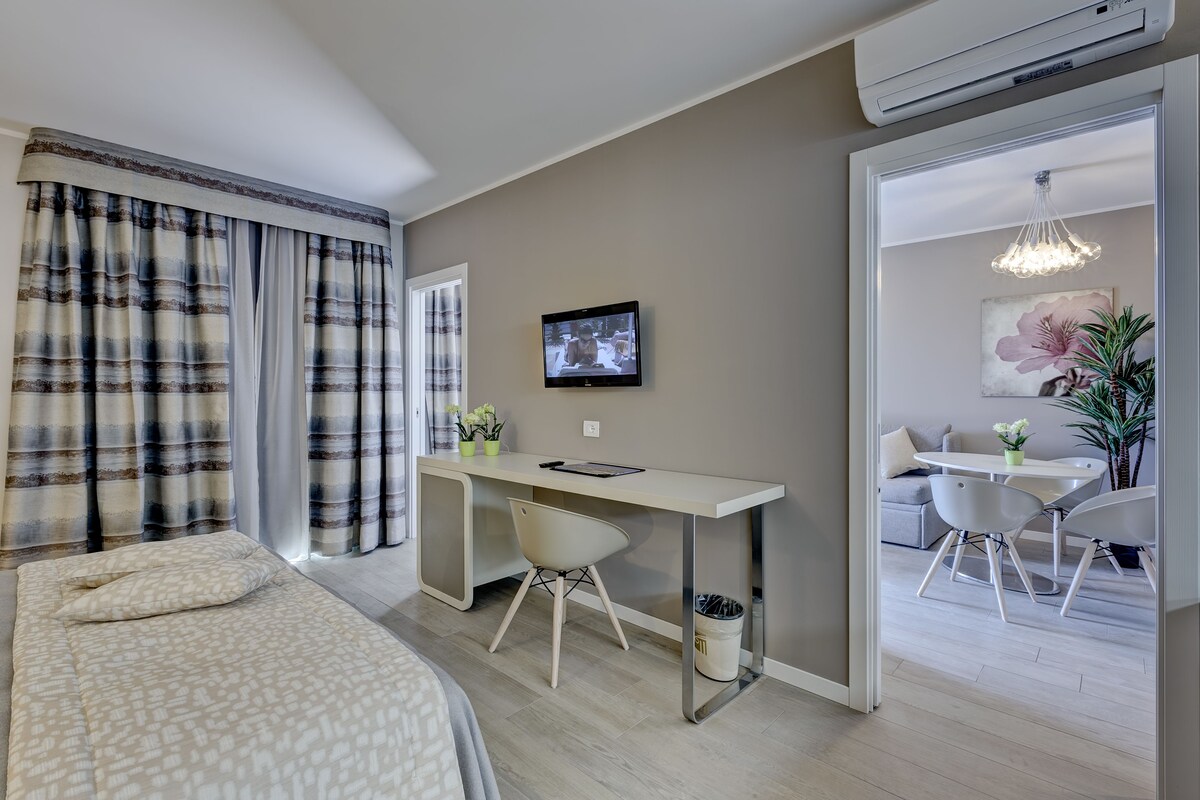 A modern hotel room with a bed, a desk with a chair, a TV, and a dining area with chairs and a table.