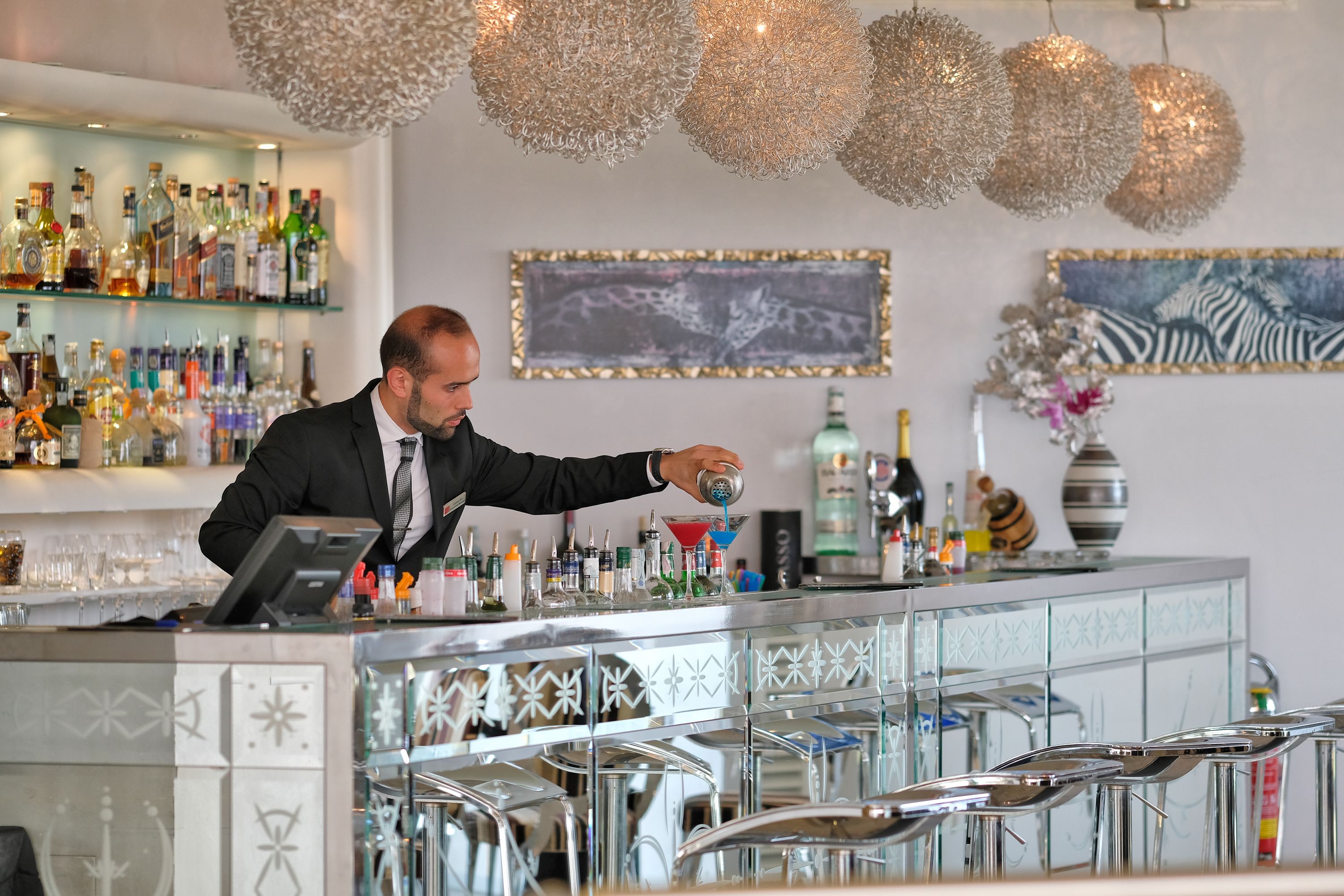 A bar area with a bartender preparing a drink, shelves with various bottles, and decorative wall art.