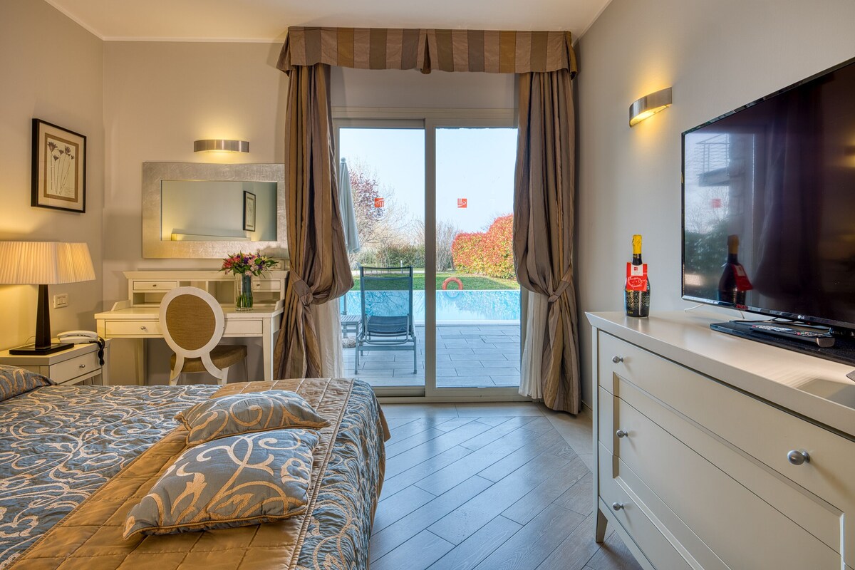 A hotel room with a bed, a desk, a TV, and a view of a garden and pool.