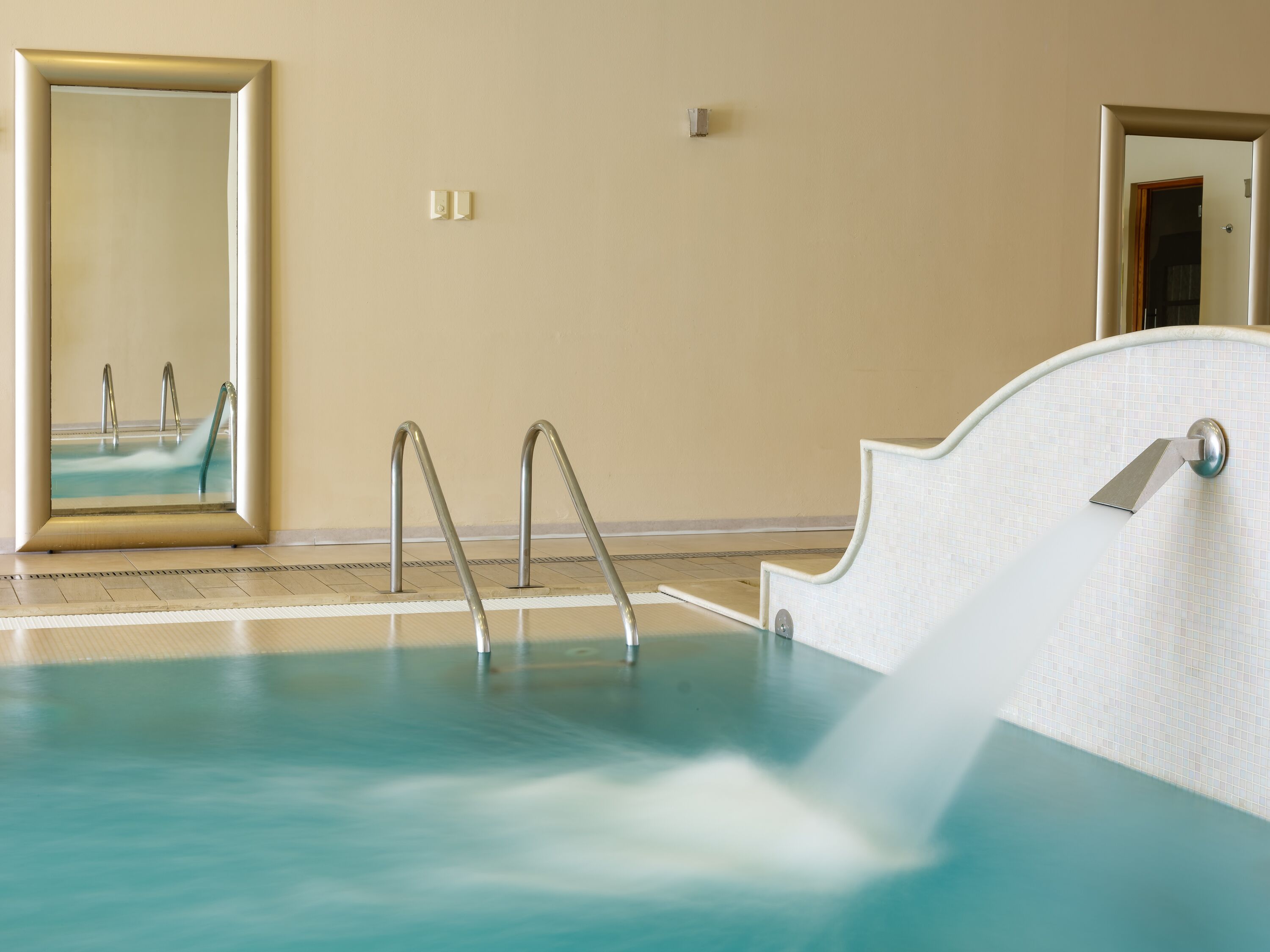 A swimming pool with metal ladders, a tiled floor, and a wall-mounted mirror.