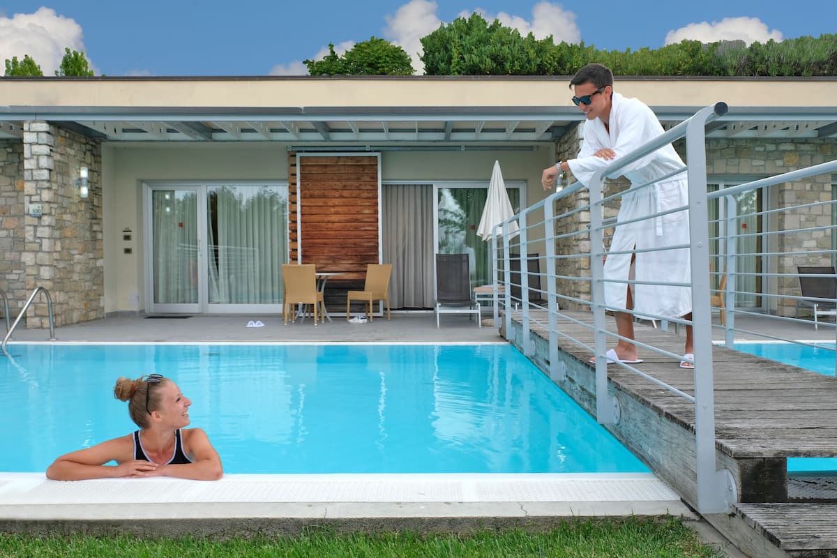 A swimming pool with a wooden deck, a stone wall, and a glass door leading to an outdoor area with a table and chairs.