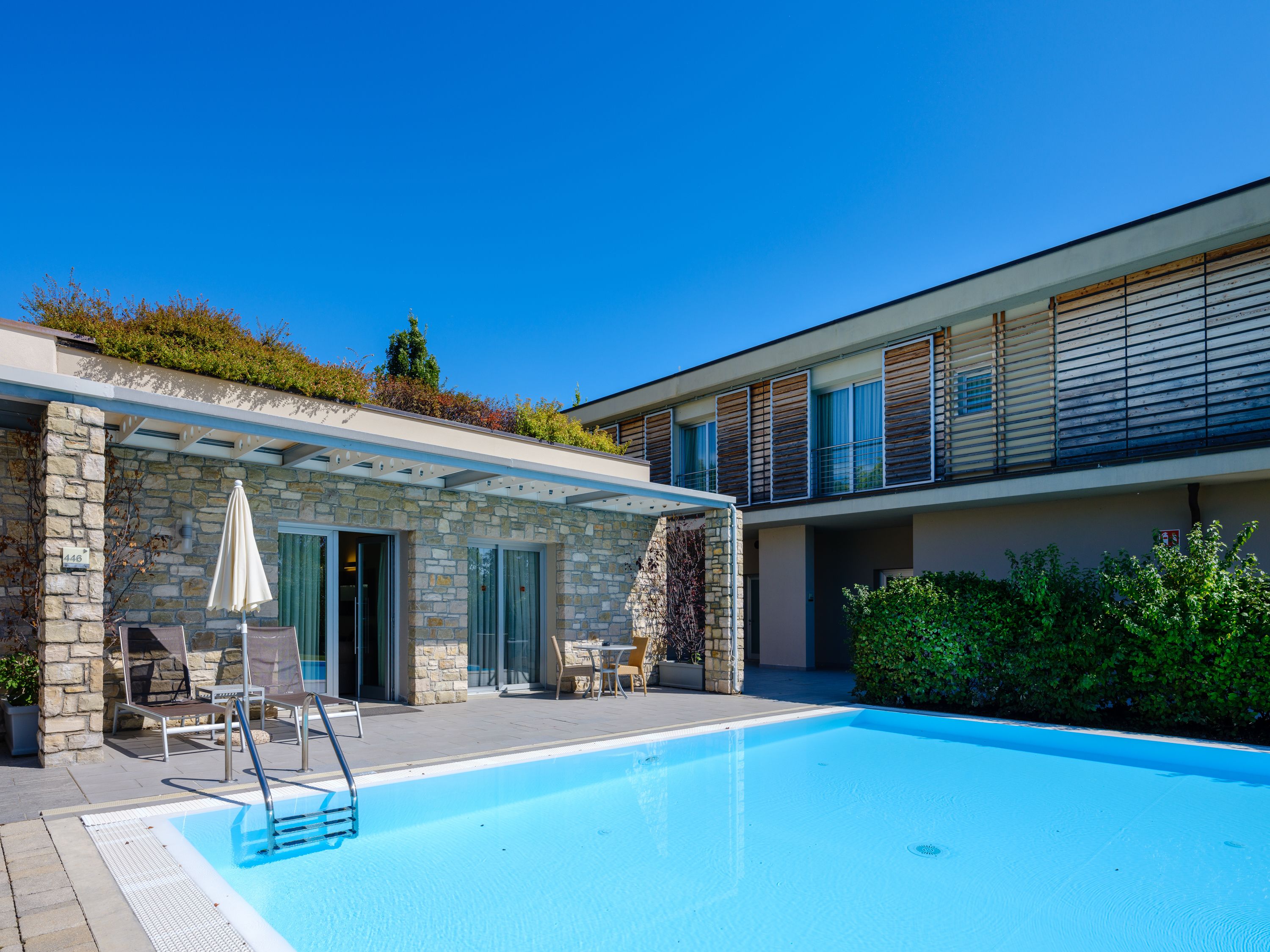 Un área moderna junto a la piscina con un muro de piedra, mobiliario exterior y una piscina azul cristalina.