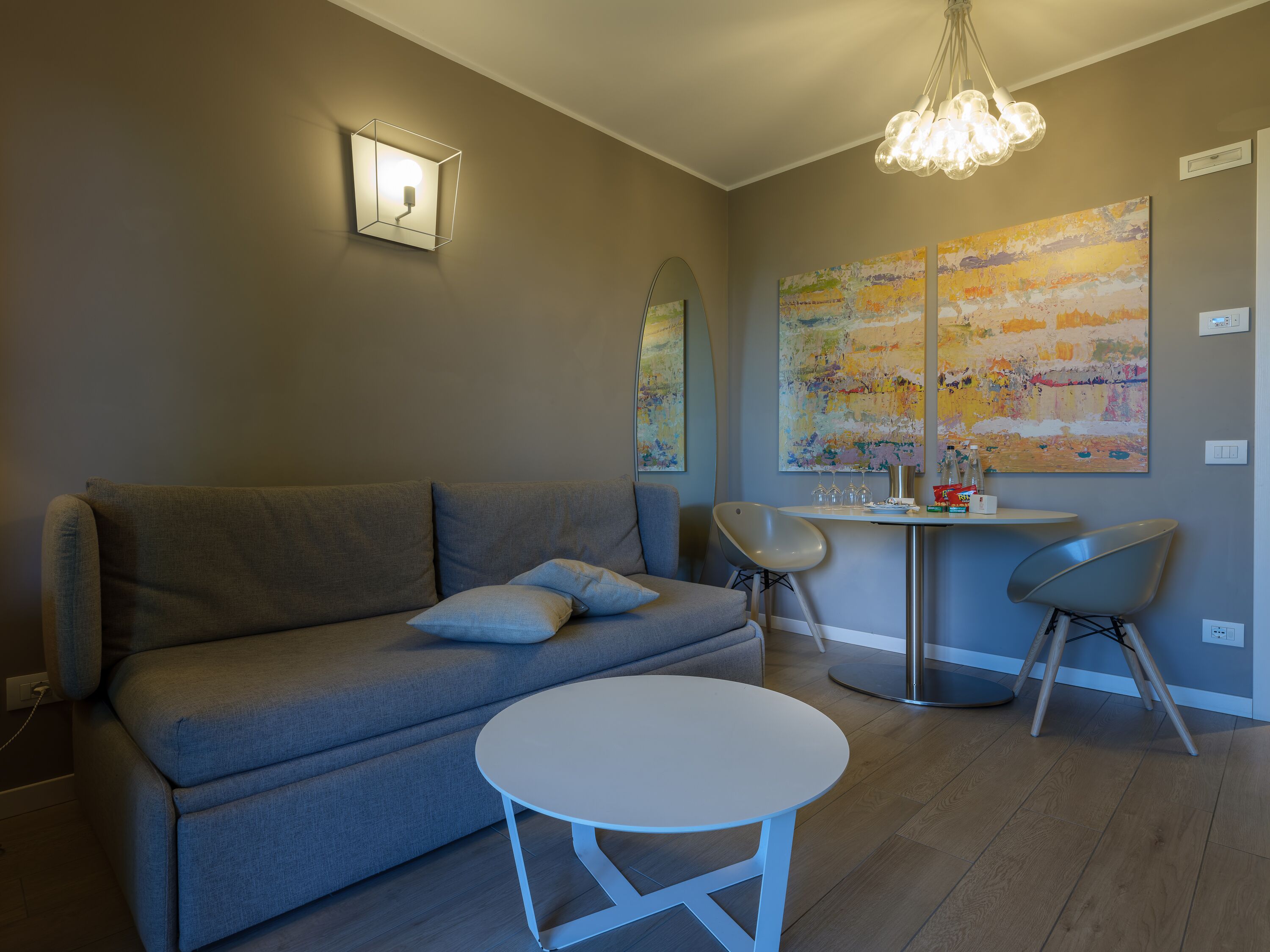 Una sala de estar moderna con un sofá gris, una mesa redonda blanca y dos sillas. Hay una araña de cristal sobre la mesa y un cuadro en la pared.