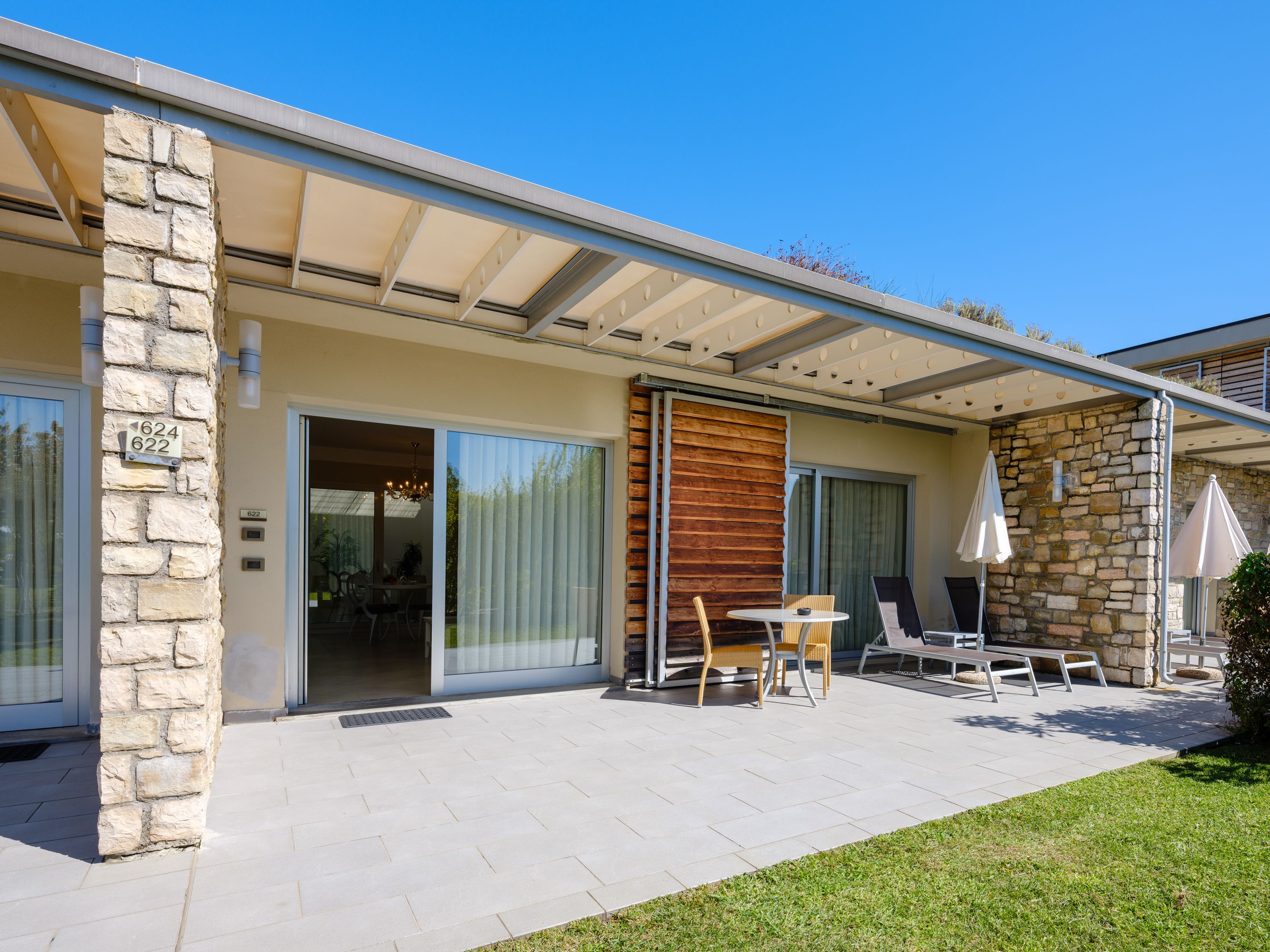 A modern house with a stone exterior, a covered patio with outdoor furniture, and a sliding glass door leading to an interior dining area.