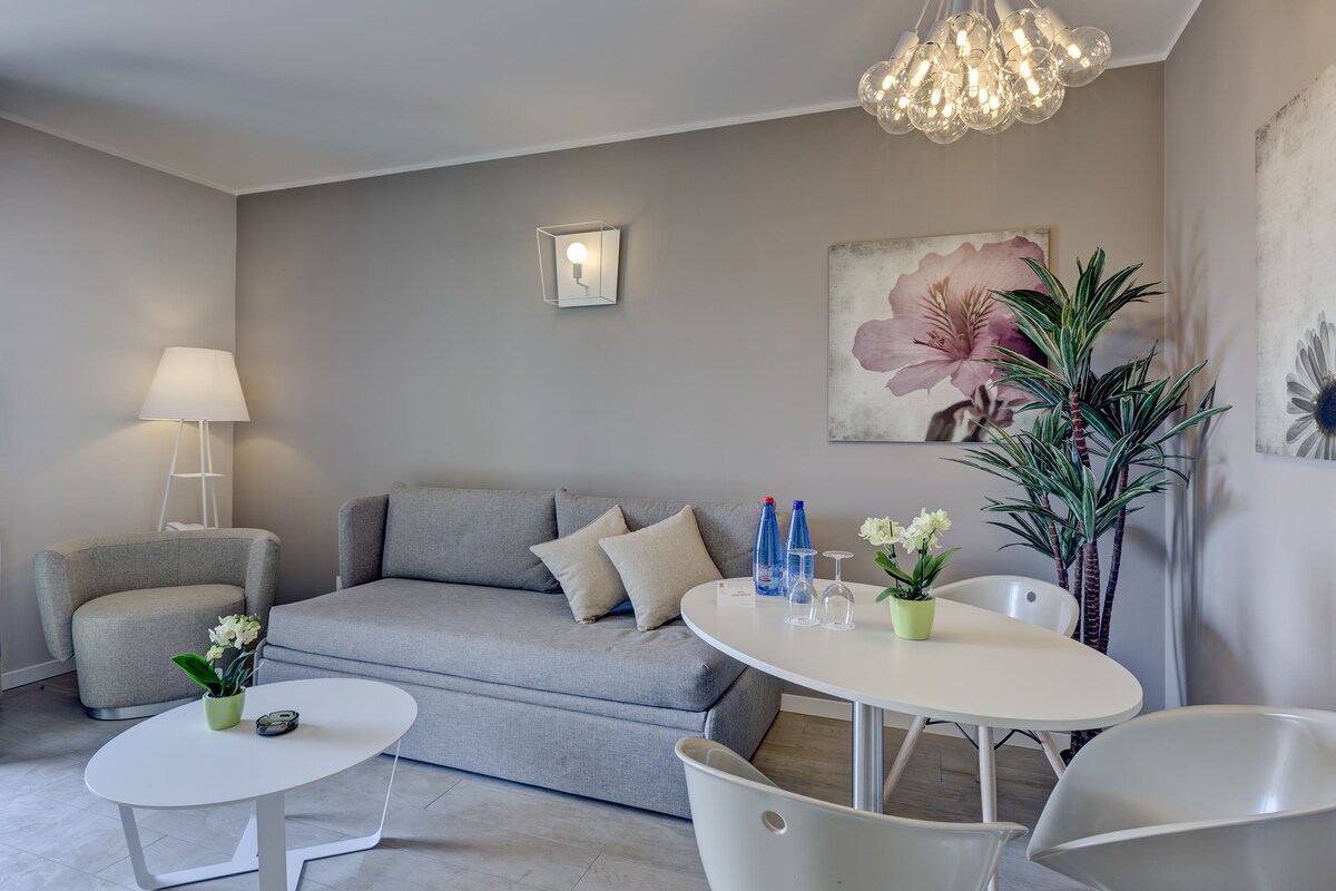 A modern living room with a grey sofa, a white coffee table, a white armchair, a lamp, and a painting on the wall.