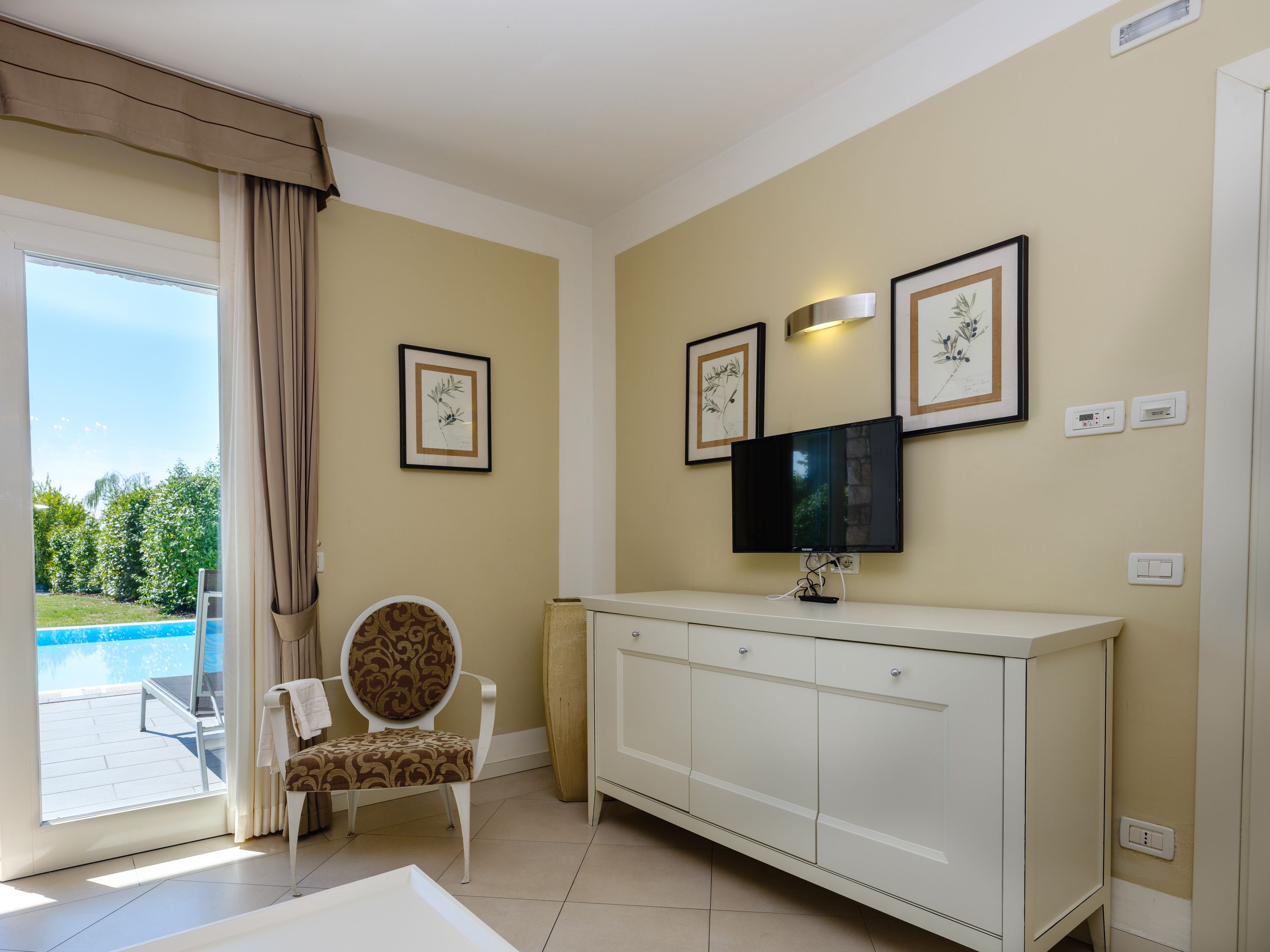 Habitación con un ventanal grande, un armario blanco, un televisor montado en la pared y una silla con cojín estampado.