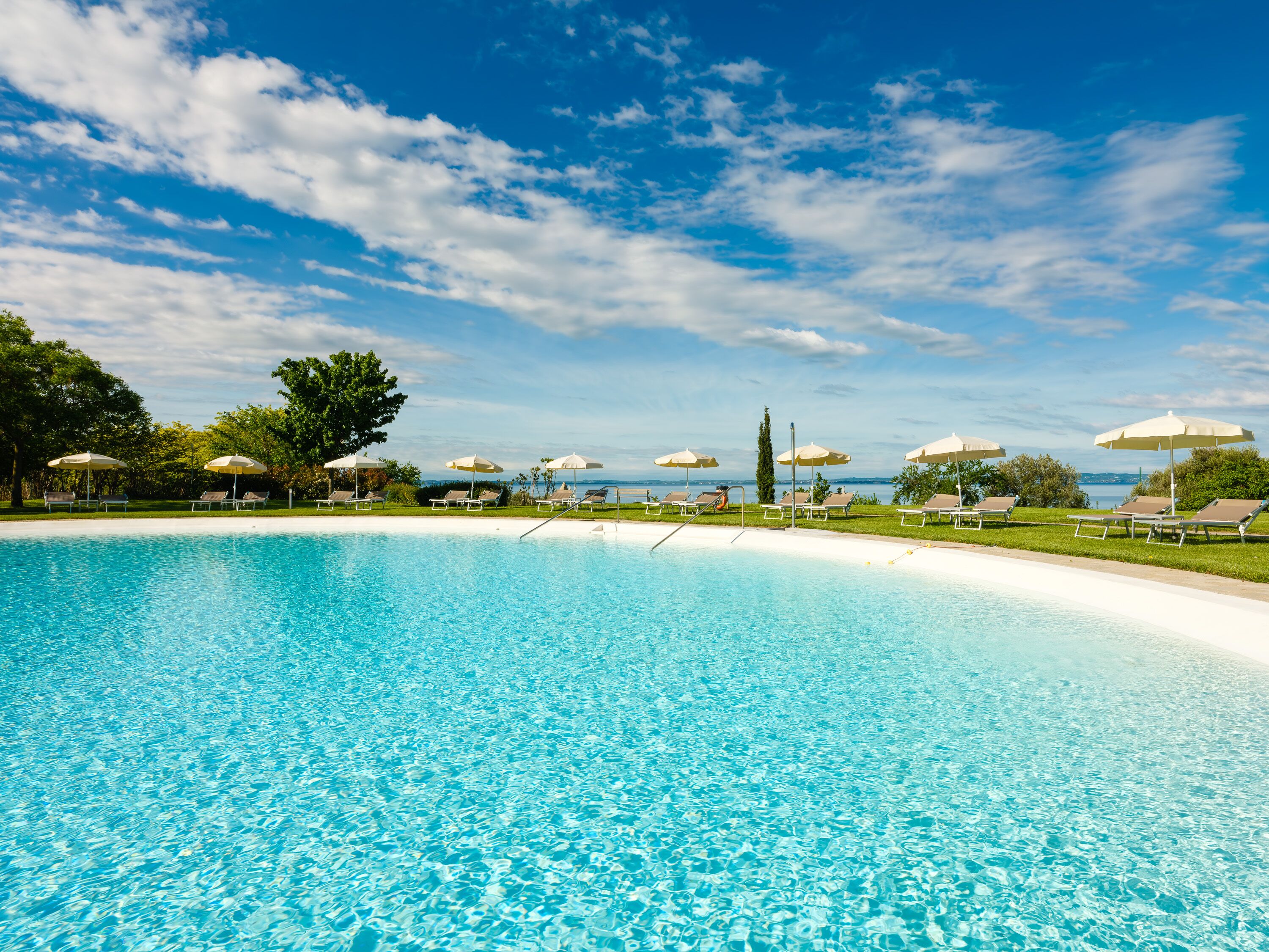 A clear blue pool with sun loungers and umbrellas, surrounded by trees and a view of the sea.