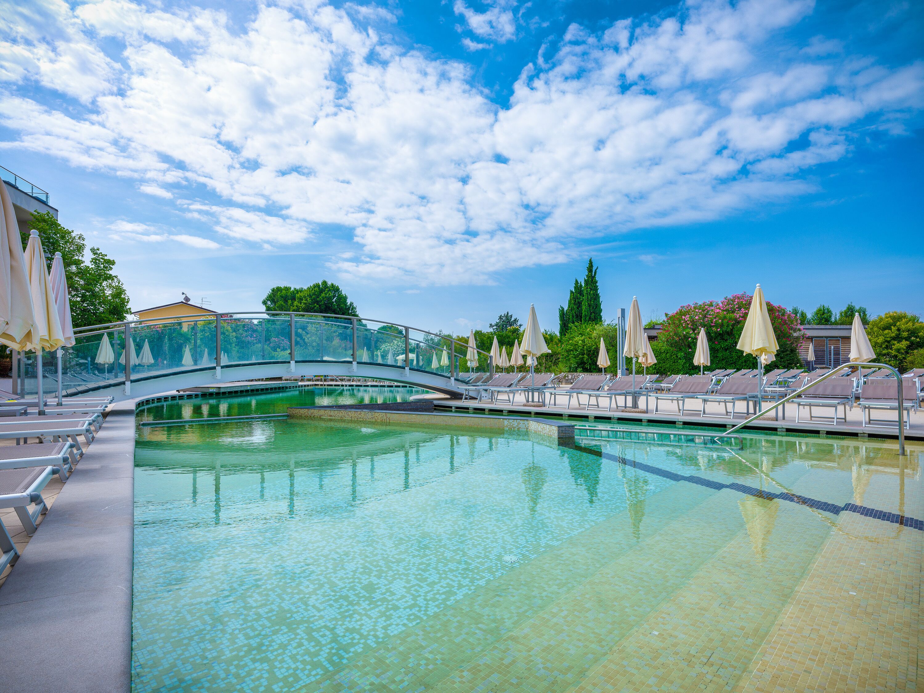 Ein Schwimmbecken mit Liegestühlen und Sonnenschirmen, eine Glasbrücke und ein strahlend blauer Himmel.