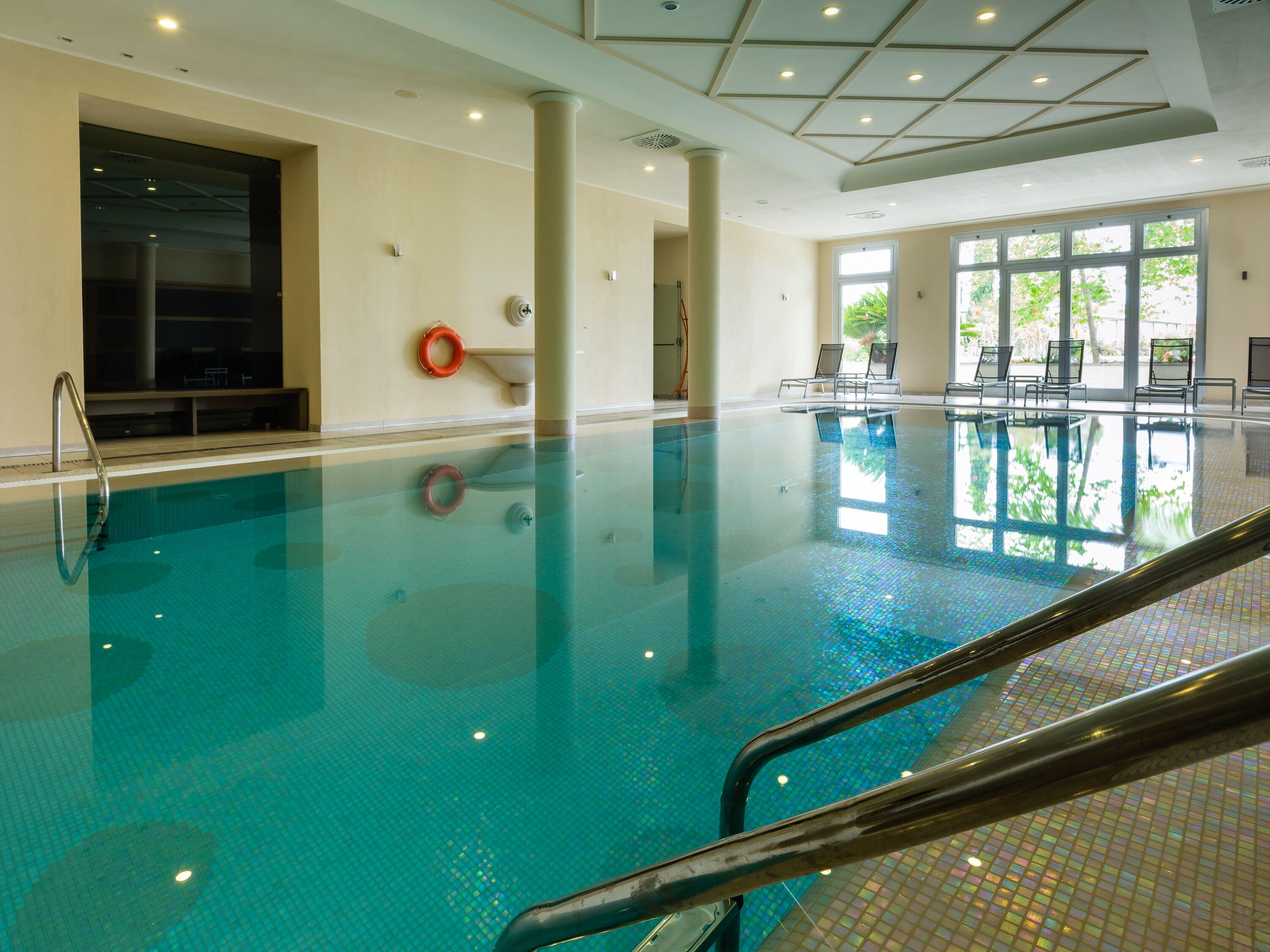 Ein Hallenbad mit klarem Wasser, einem Rettungsschwimmerstuhl und einem sonnendurchfluteten Bereich mit Stühlen und einem Tisch.