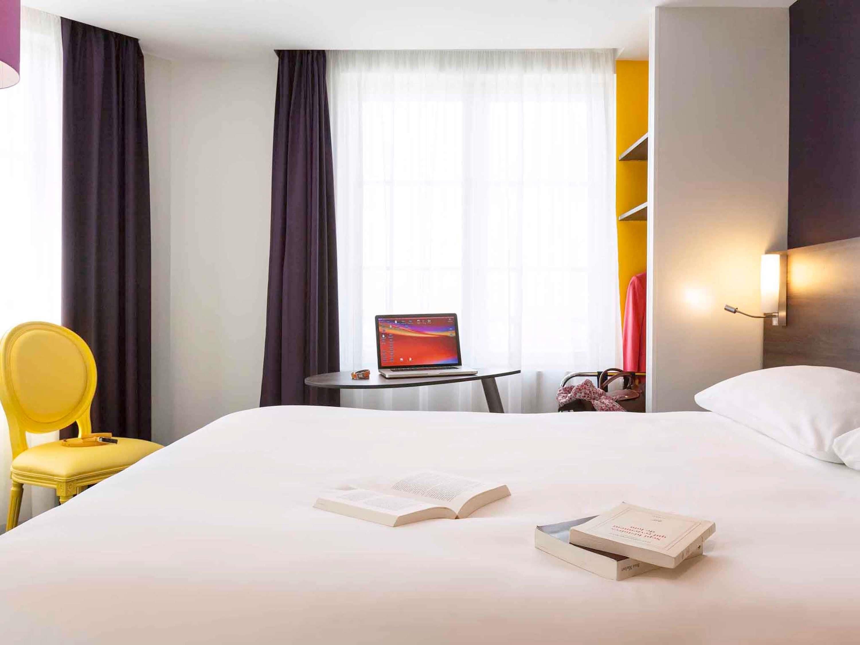 A hotel room with a bed, a laptop on a small table, a chair, and a bedside table with a lamp.