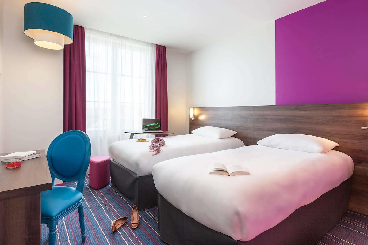 A hotel room with two beds, a desk with a laptop, a chair, and a window with curtains.