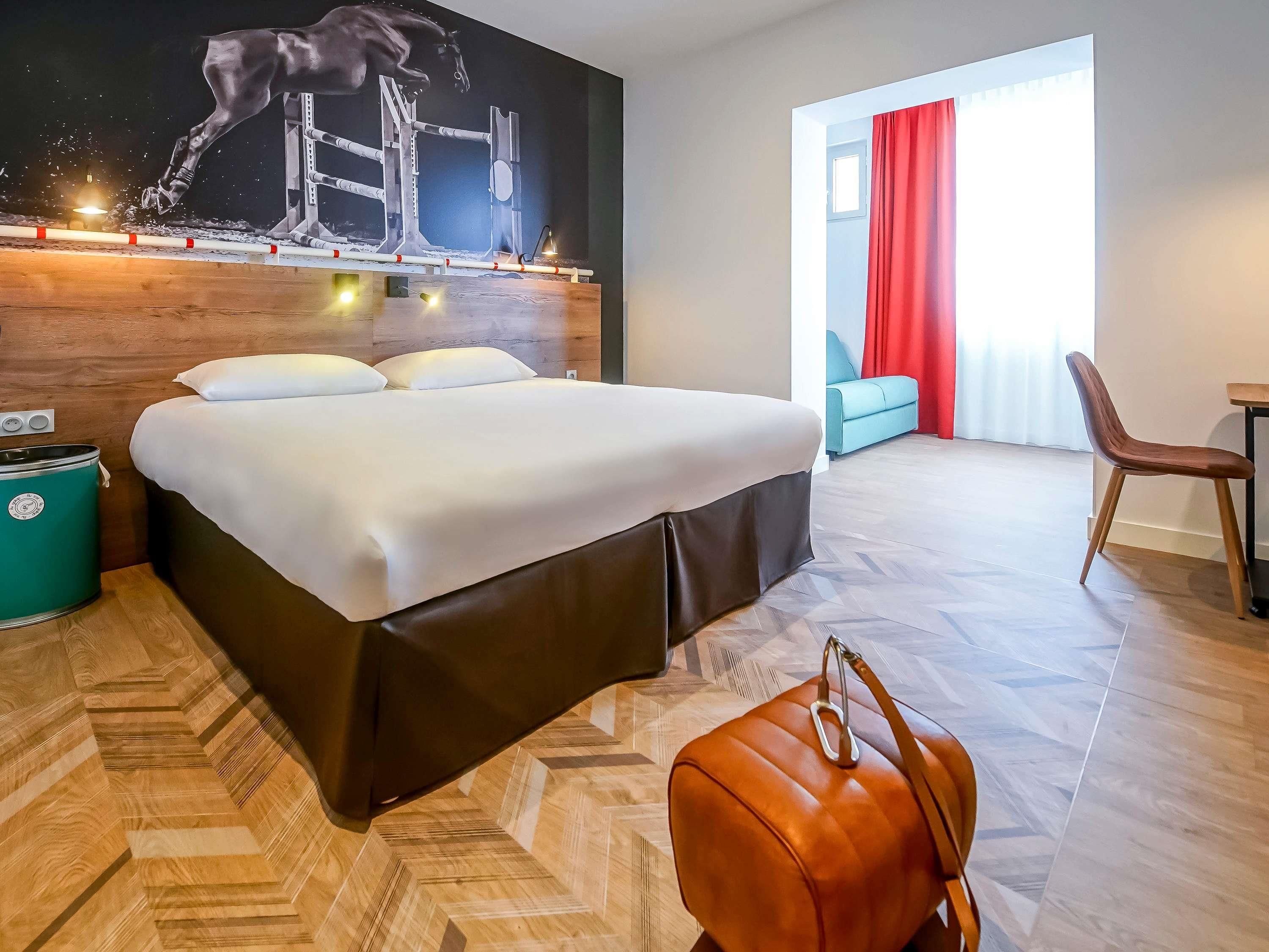 A hotel room with a bed, a painting, a suitcase, and a view of the outside.