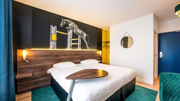 A modern hotel room with a large bed, a wooden headboard, a wall-mounted horse jumping sculpture, a round mirror, and a green chair.