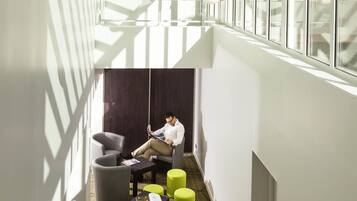 A modern hotel lobby with a man reading a magazine, a seating area with a sofa and ottomans, and large windows allowing natural light.