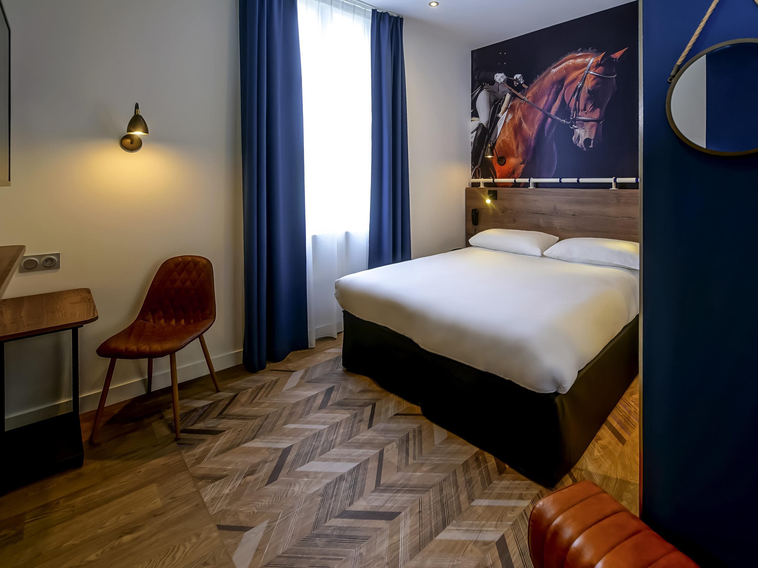 A hotel room with a bed, a chair, a desk, and a large window with curtains.