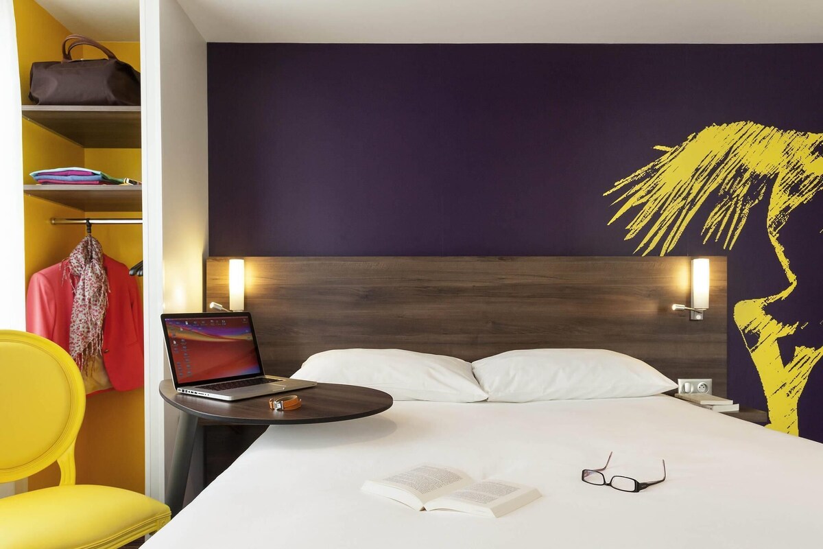 Una habitación de hotel con una cama, un ordenador portátil sobre una mesita de noche, un libro y gafas.