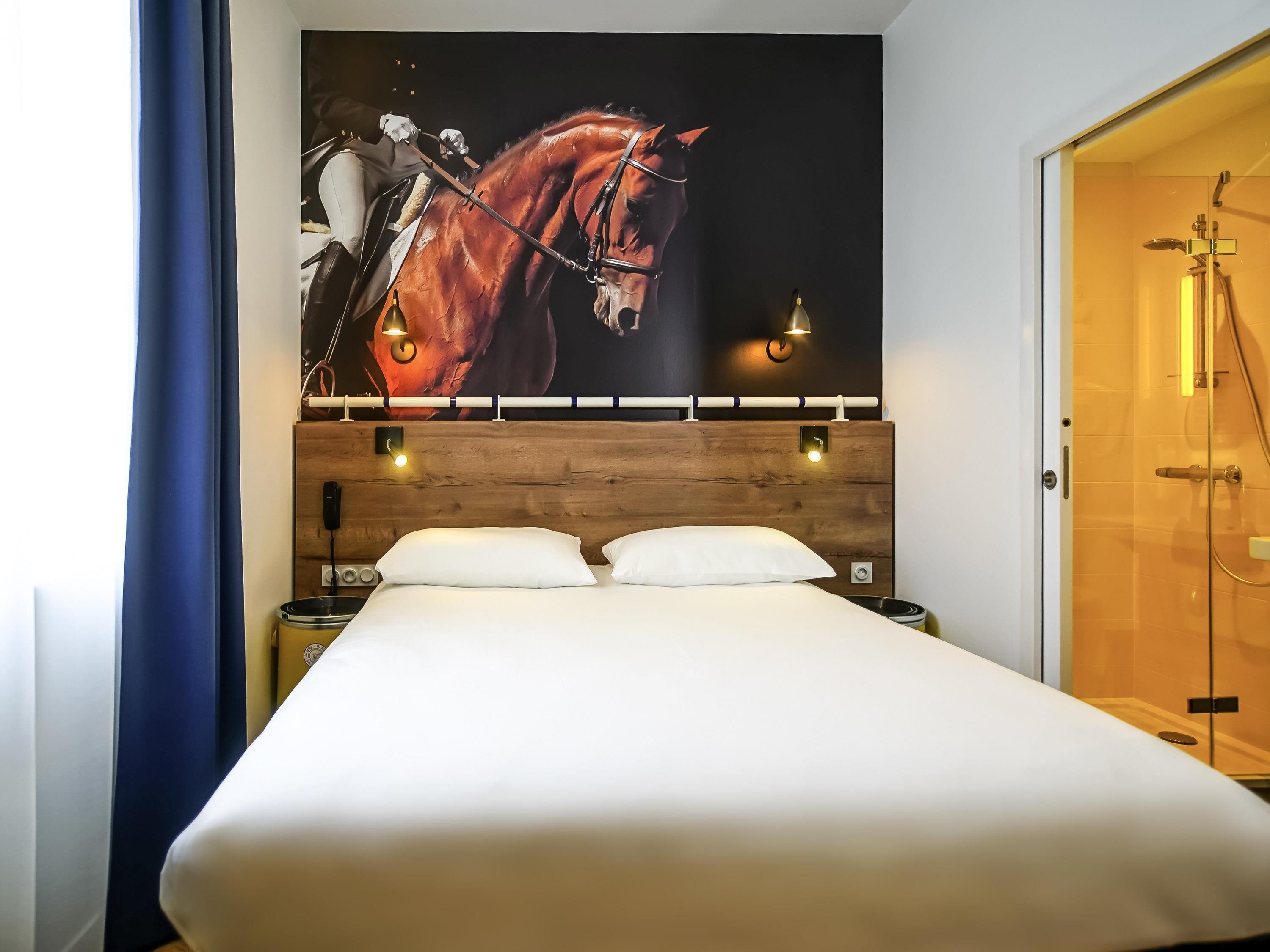 Habitación de hotel con una cama grande, cabecero de madera, dos mesitas de noche con lámparas y un cuadro grande de un caballo en la pared.