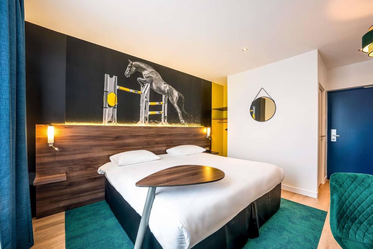 Une chambre d’hôtel moderne avec un grand lit, une tête de lit en bois, une sculpture murale représentant un cheval sautant, un miroir rond et un fauteuil vert.