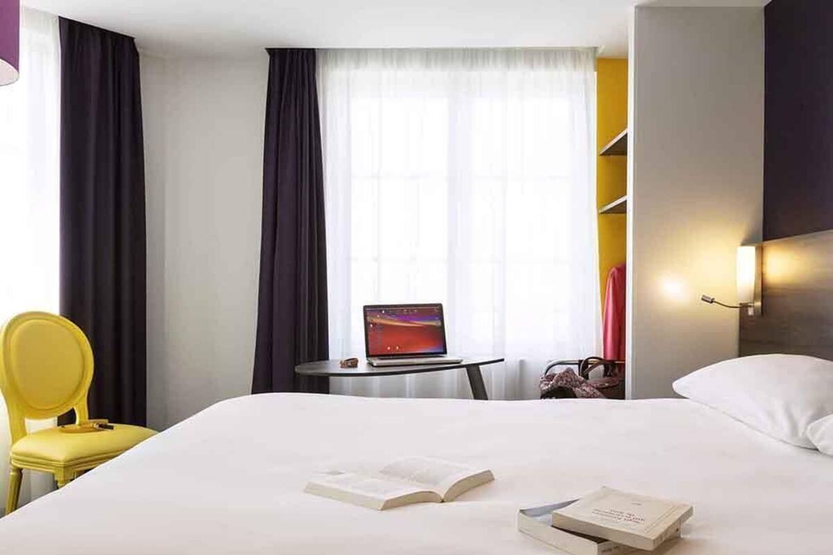Une chambre d’hôtel avec un lit, un ordinateur portable posé sur une petite table, une chaise et une fenêtre avec des rideaux.
