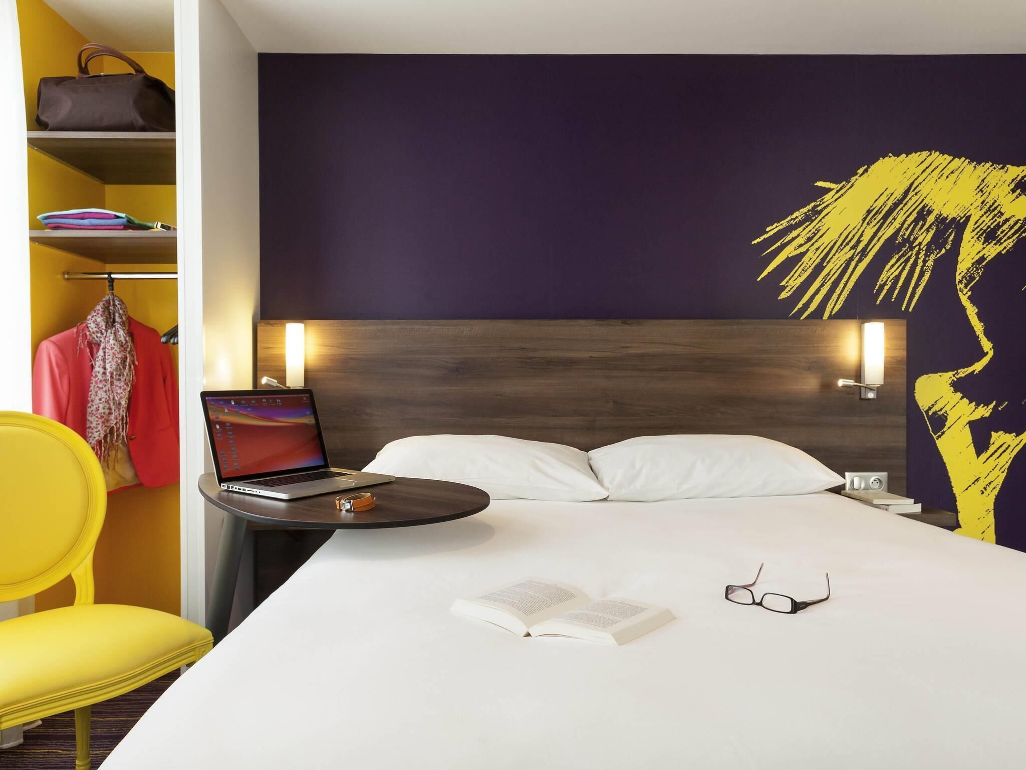Een hotelkamer met een bed, een laptop op een nachtkastje, een boek en een bril.