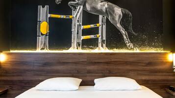 Una habitación con un cuadro grande de un caballo saltando un obstáculo, un cabecero de madera y dos almohadas blancas sobre la cama.