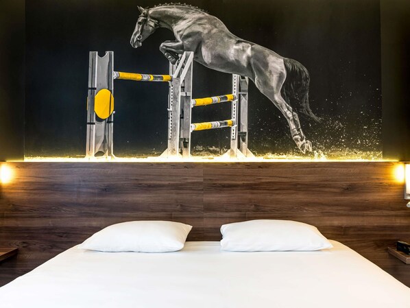 Una habitación con un cuadro grande de un caballo saltando un obstáculo, un cabecero de madera y dos almohadas blancas sobre la cama.