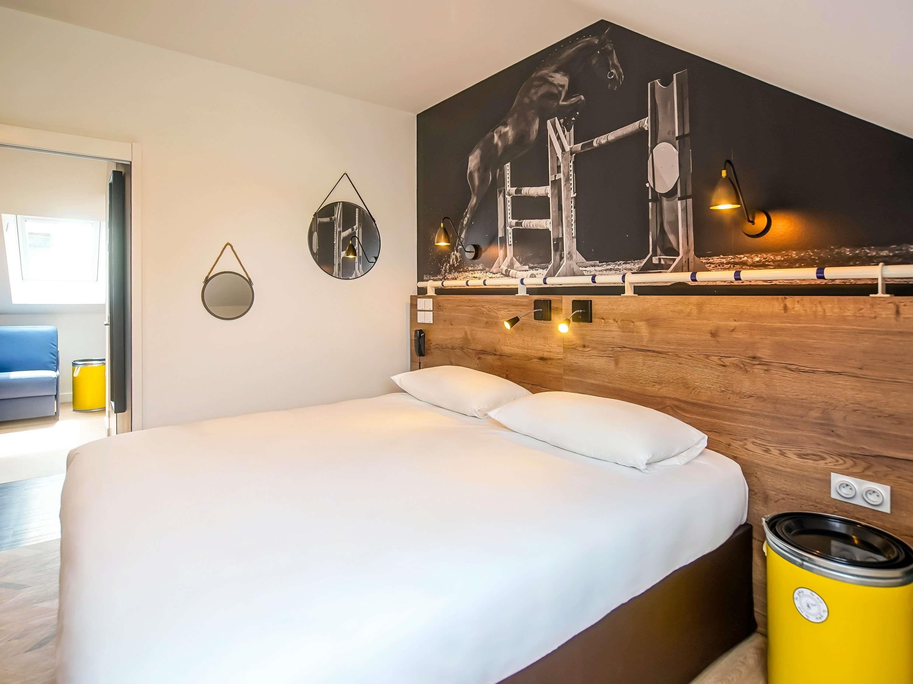 Een hotelkamer met een groot bed, een houten hoofdeinde, een schilderij van een paard aan de muur en een gele prullenbak.