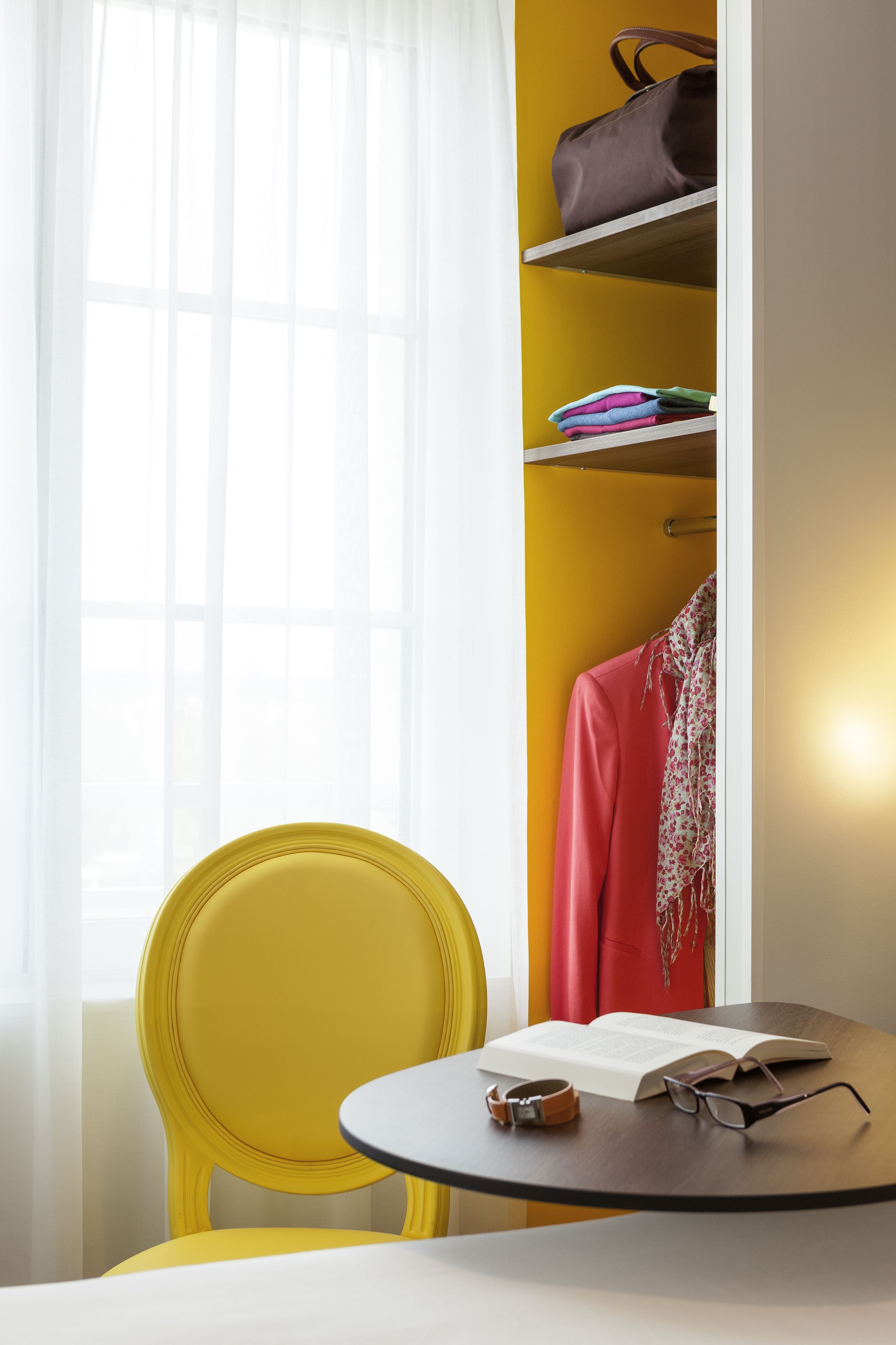A room with a yellow chair, a table with a book and glasses, and a wardrobe with folded clothes and a handbag.
