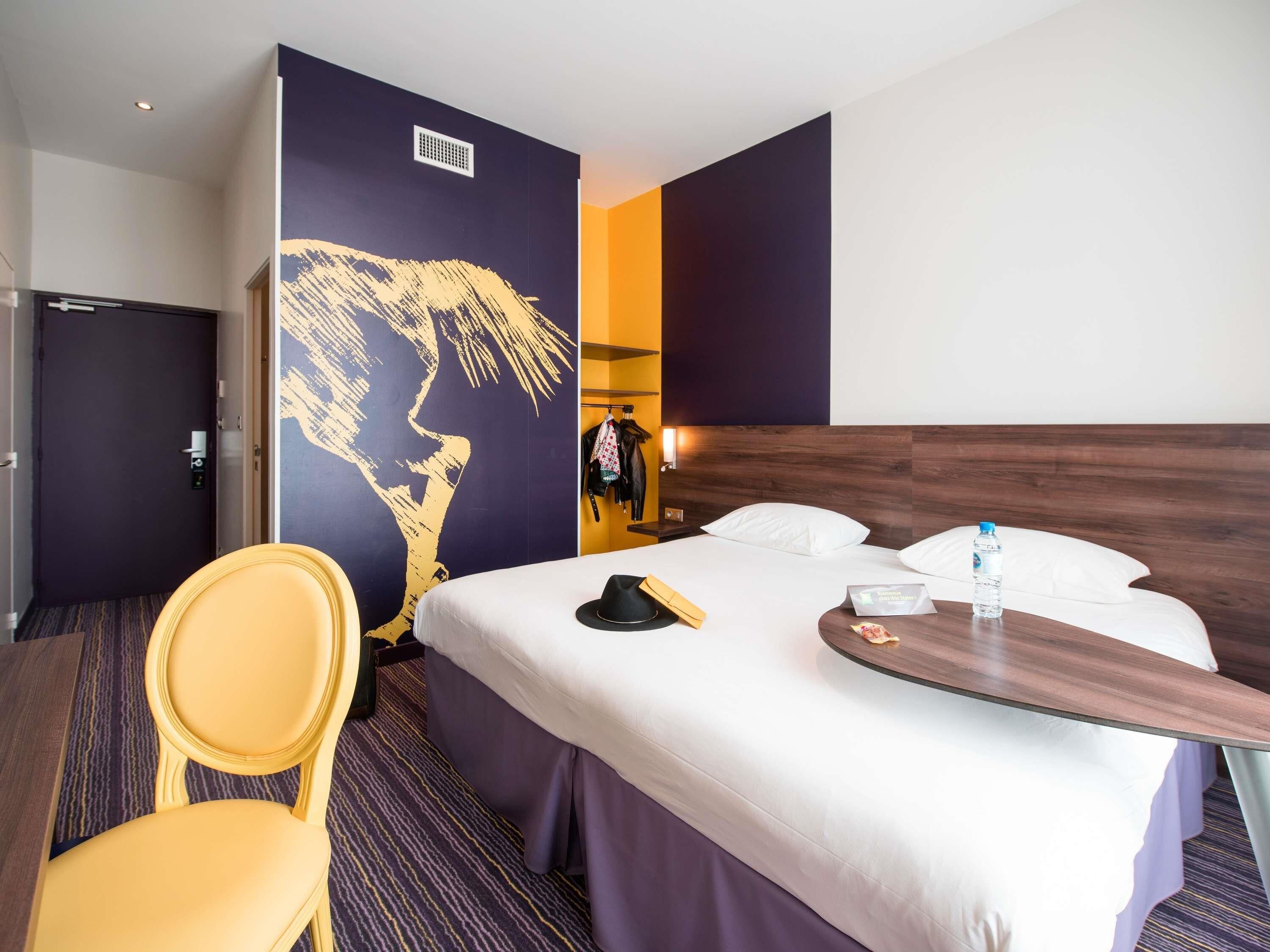 Une chambre d’hôtel avec un grand lit, une chaise, une tête de lit en bois et un mur orné d’un grand tableau abstrait.