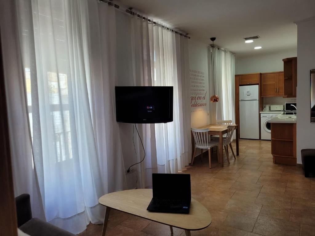 A living room with a flat-screen TV, a laptop on a table, a dining area with chairs, and a kitchen with cabinets and appliances.