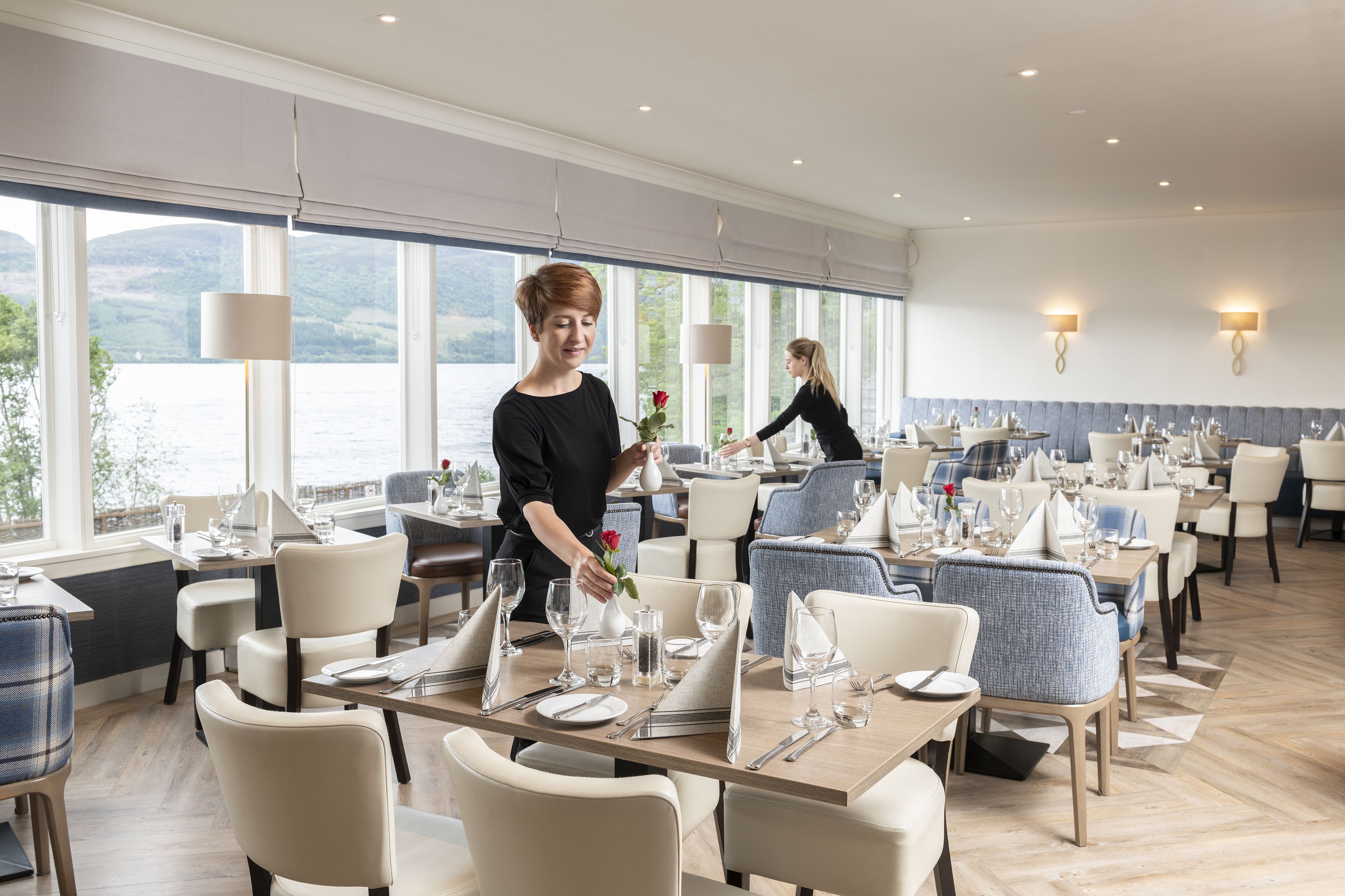 Un restaurant où les tables sont dressées pour le service, de grandes fenêtres offrant une vue sur l’extérieur, et un serveur s’occupant d’une table.