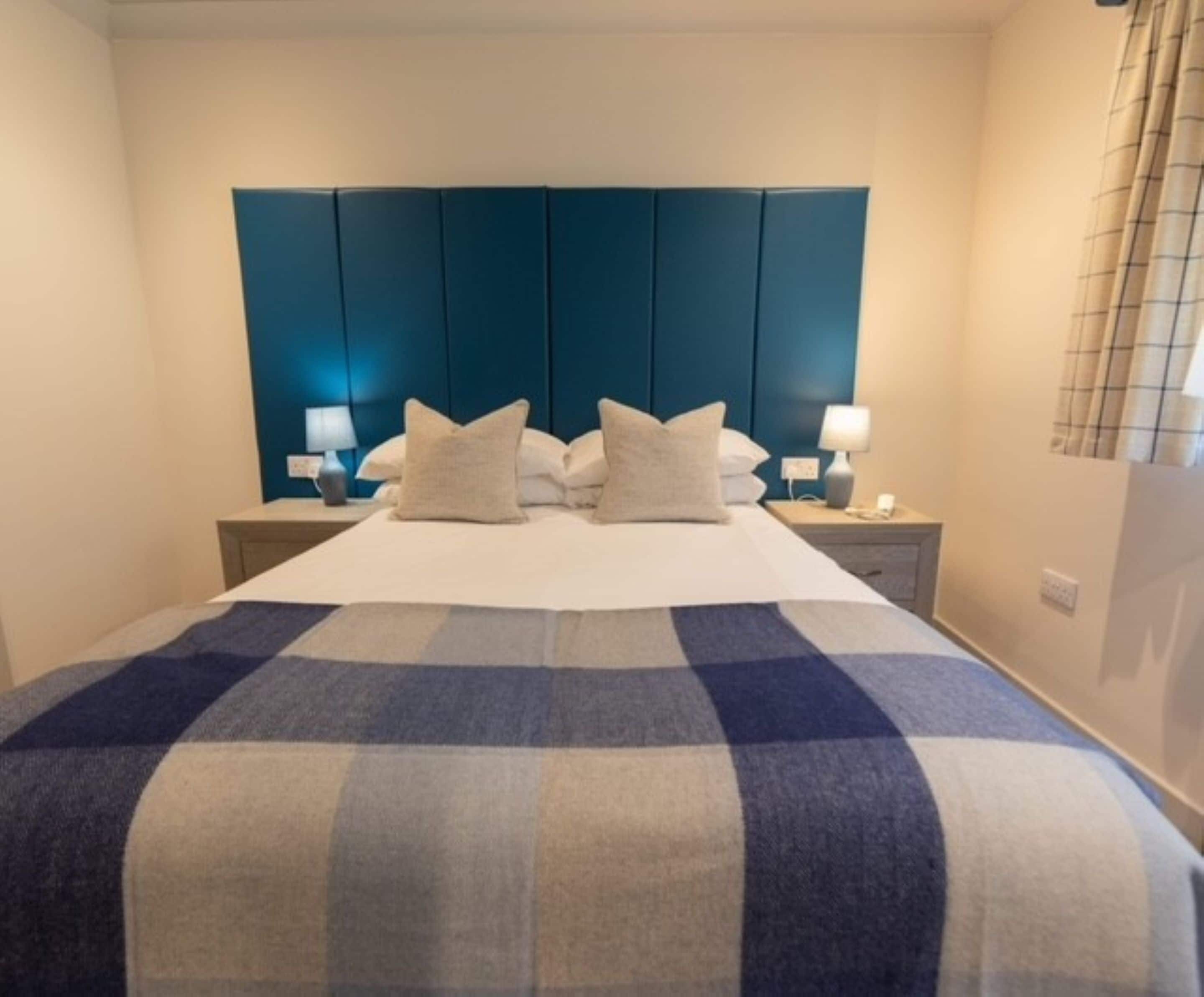 Un lit bien fait, avec une couverture à carreaux, une tête de lit bleue et des lampes de chevet.