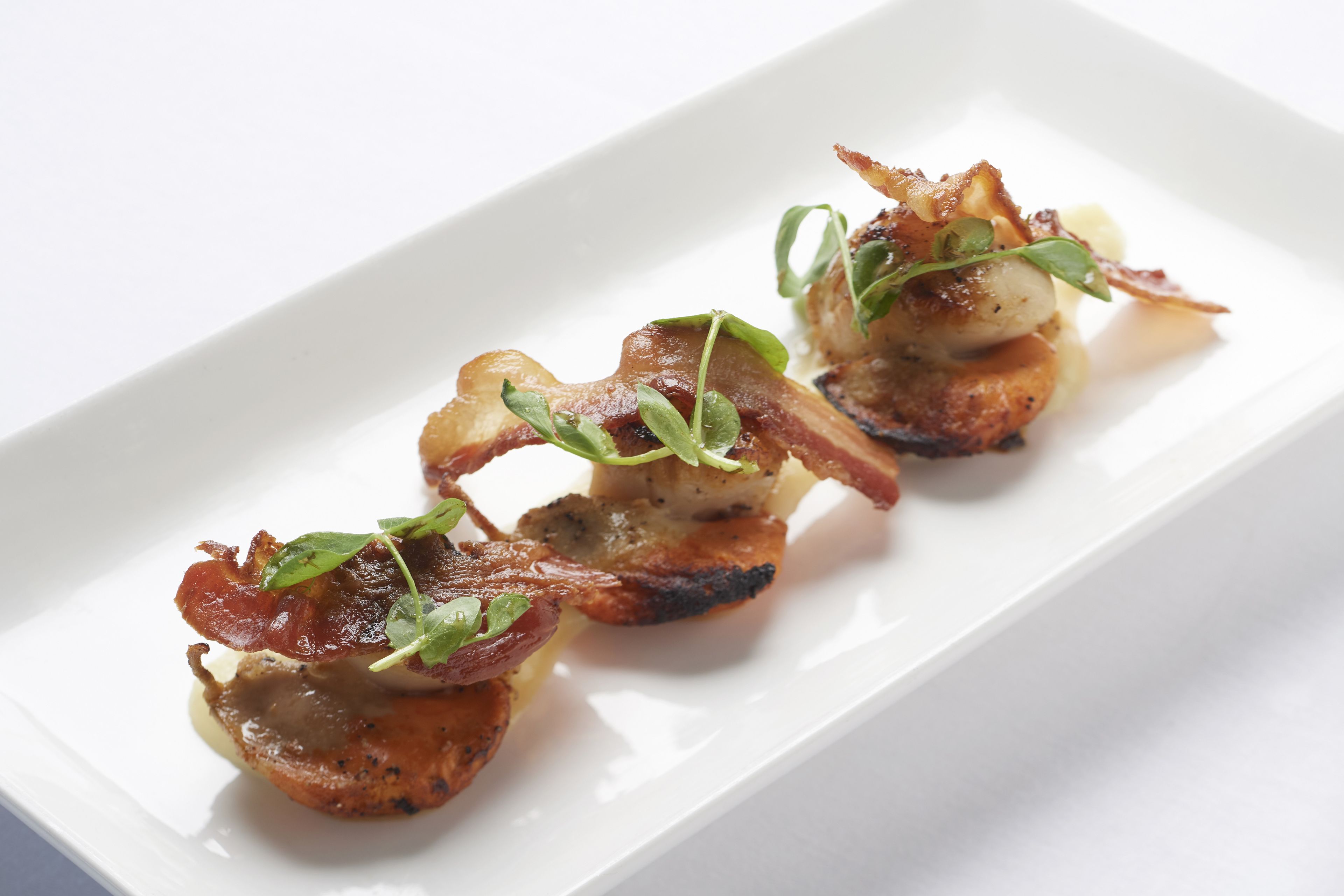 A white rectangular plate with three pieces of food, each topped with bacon and garnished with green leaves.