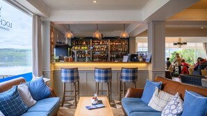 Zona de bar con vista al lago, un espacio de estar con sofá y sillones, y una barra con diferentes botellas.