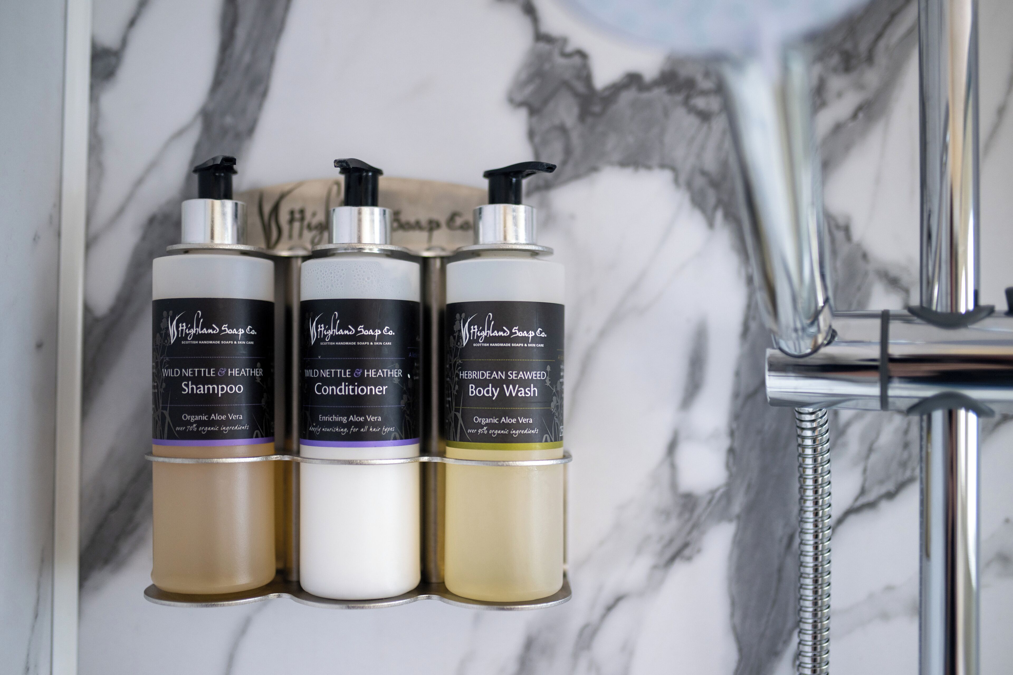Tres botellas de champú, acondicionador y jabón de cuerpo Wild Nettle & Heather sobre una pared de mármol.