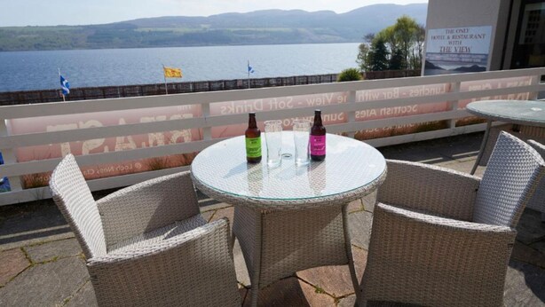 Una mesa de vidrio con dos sillas de mimbre, dos botellas de cerveza y vista a un lago con montañas al fondo.