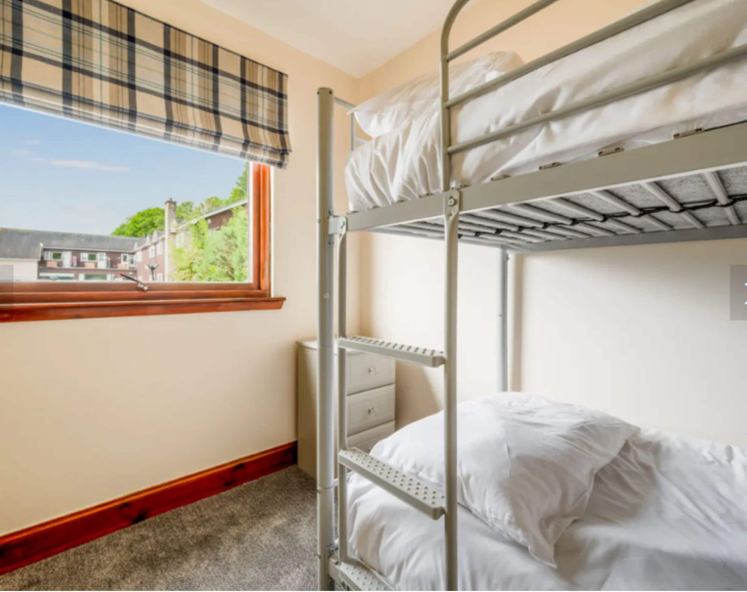Quarto com beliche, janela e vista para edifícios, com piso de madeira.
